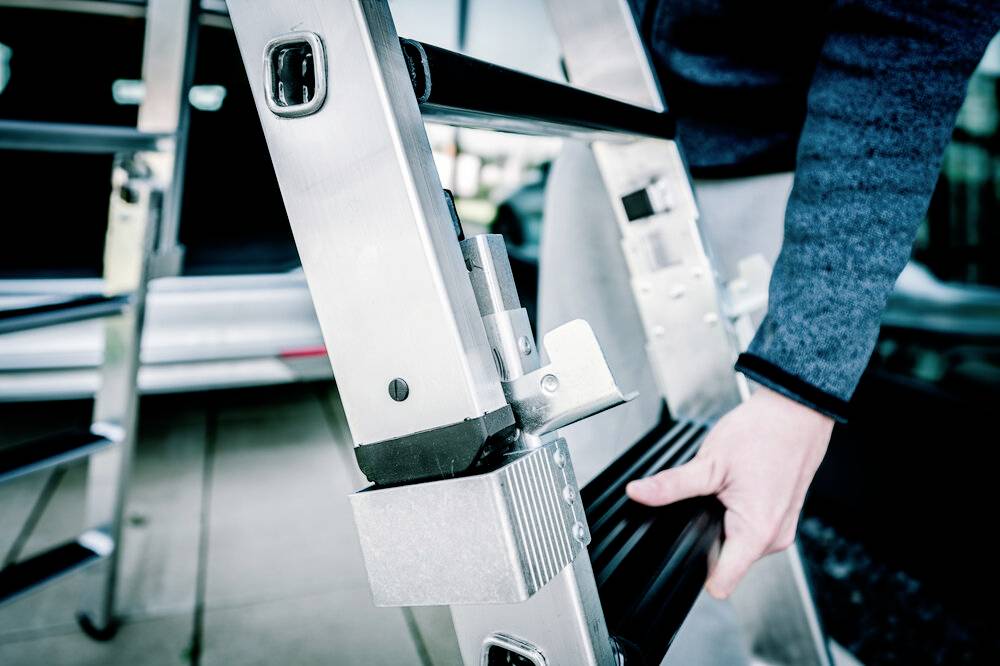 Osoba składa aluminiową drabinę. W tle widać rozmyte szczegóły samochodu.