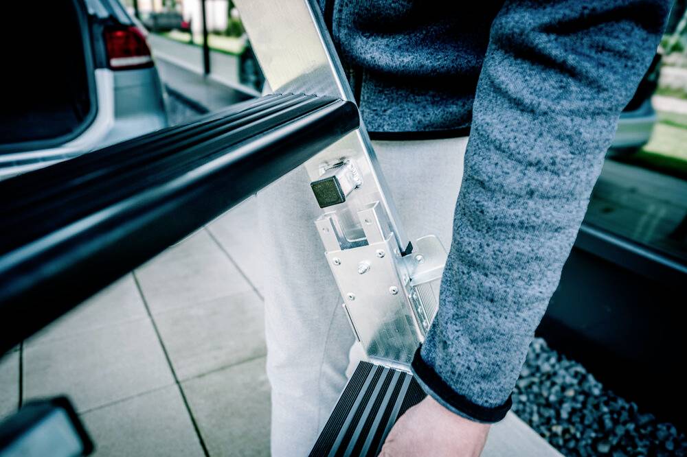 Jedna osoba trzyma metalową drabinę. W tle zaparkowany jest samochód i widać drogę.