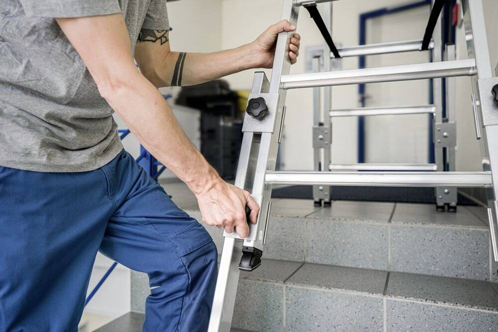 Osoba ubrana w strój roboczy ustawia aluminiową drabinę na schodach. Jej ręce regulują nogi drabiny, aby zapewnić stabilność.