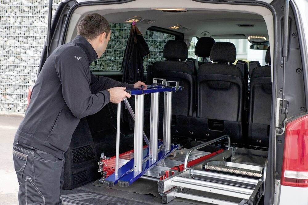 Osoba ładuje niebieską i srebrną rampę do bagażnika vana, prawdopodobnie przeznaczoną do transportu wózków inwalidzkich.