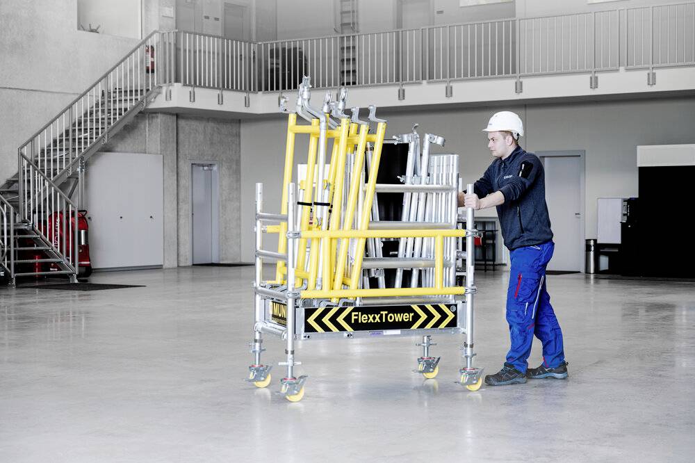 Pracownik w odzieży ochronnej przesuwa żółte rusztowanie FlexxTower na kółkach przez jasną, przestronną halę.