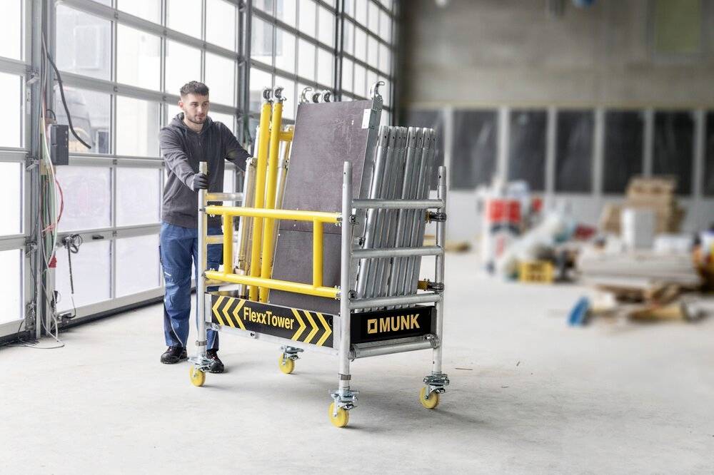 Mężczyzna przesuwa mobilną pomoc montażową z etykietą 'FlexxTower' w jasnej hali produkcyjnej. W tle widoczne są materiały budowlane.