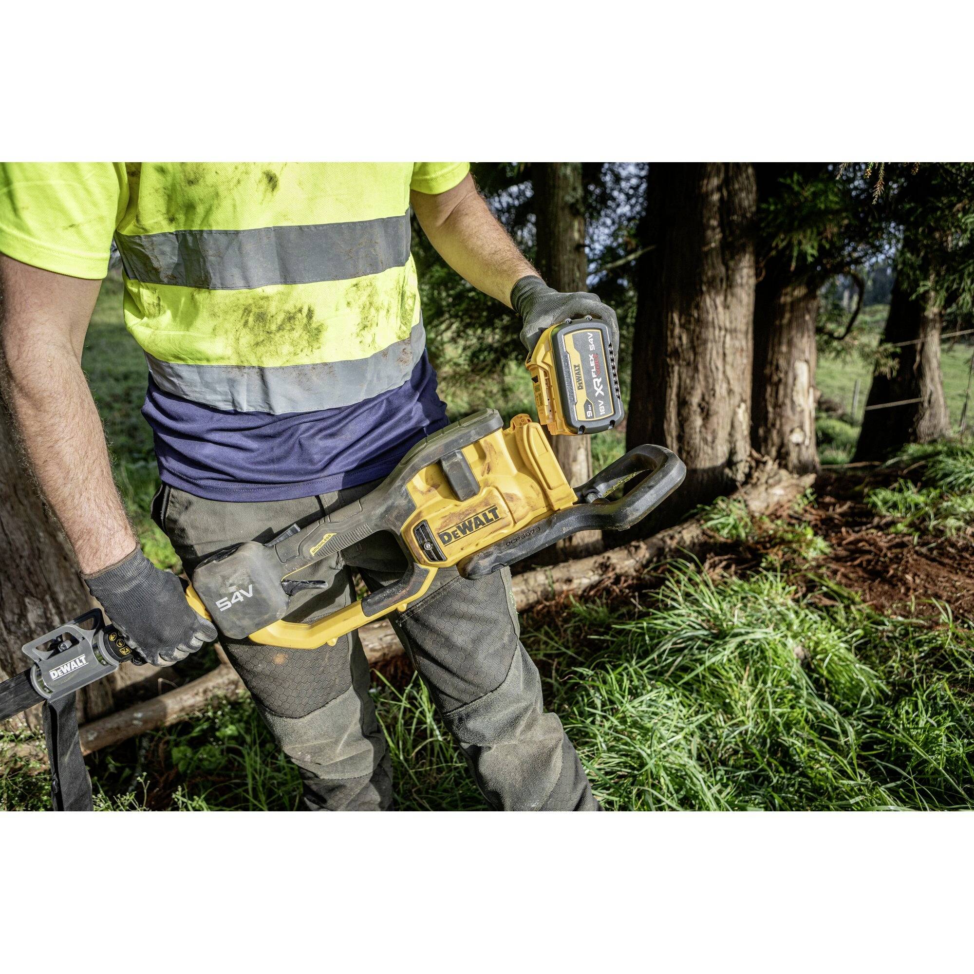 Mężczyzna w odzieży roboczej trzyma piłę łańcuchową w lesie. Ma na sobie rękawice i żółtą koszulę ochronną. W tle widoczne są drzewa.