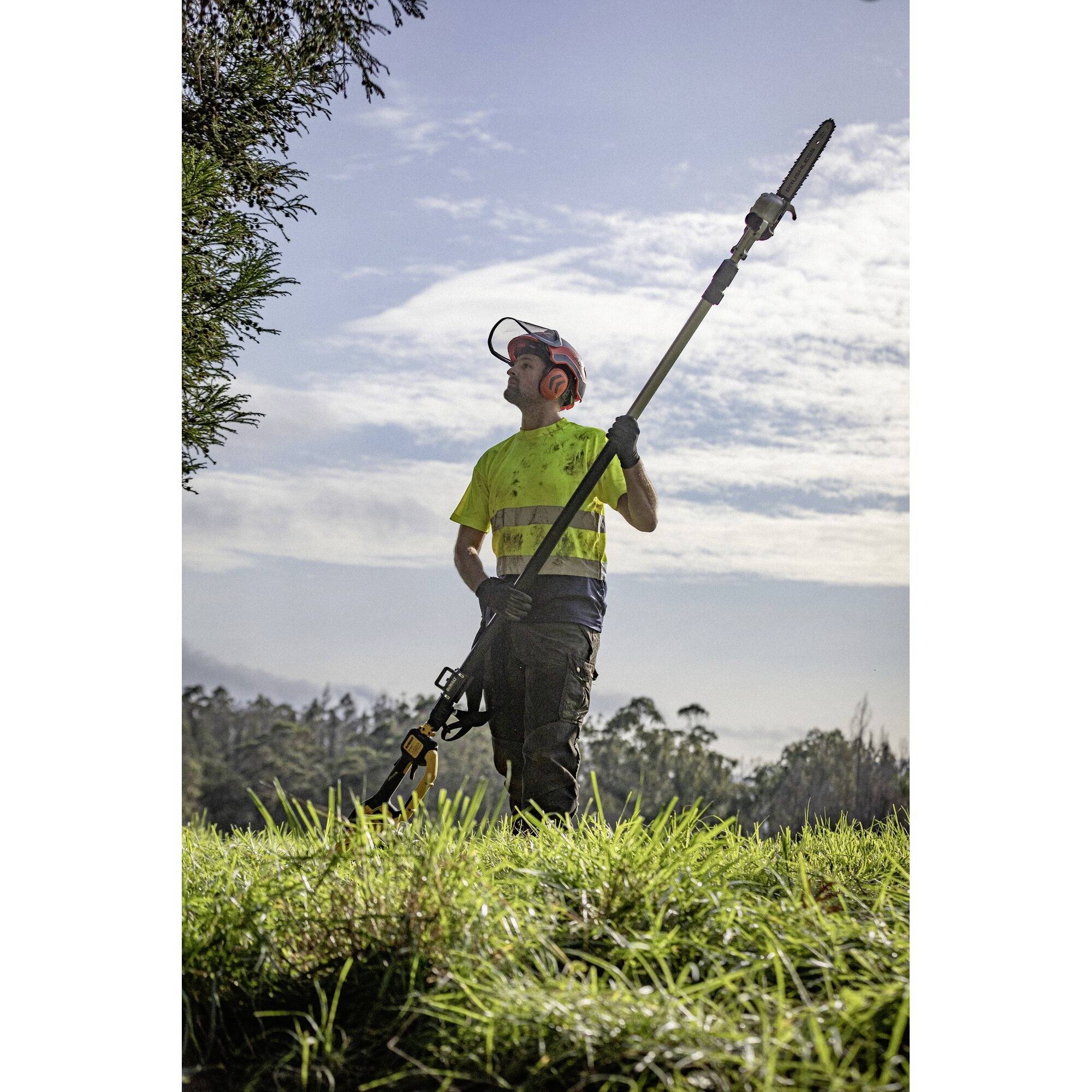 Mężczyzna nosi odzież ochronną i trzyma teleskopowe nożyce do żywopłotu. Stoi na łące przed pochmurnym niebem.