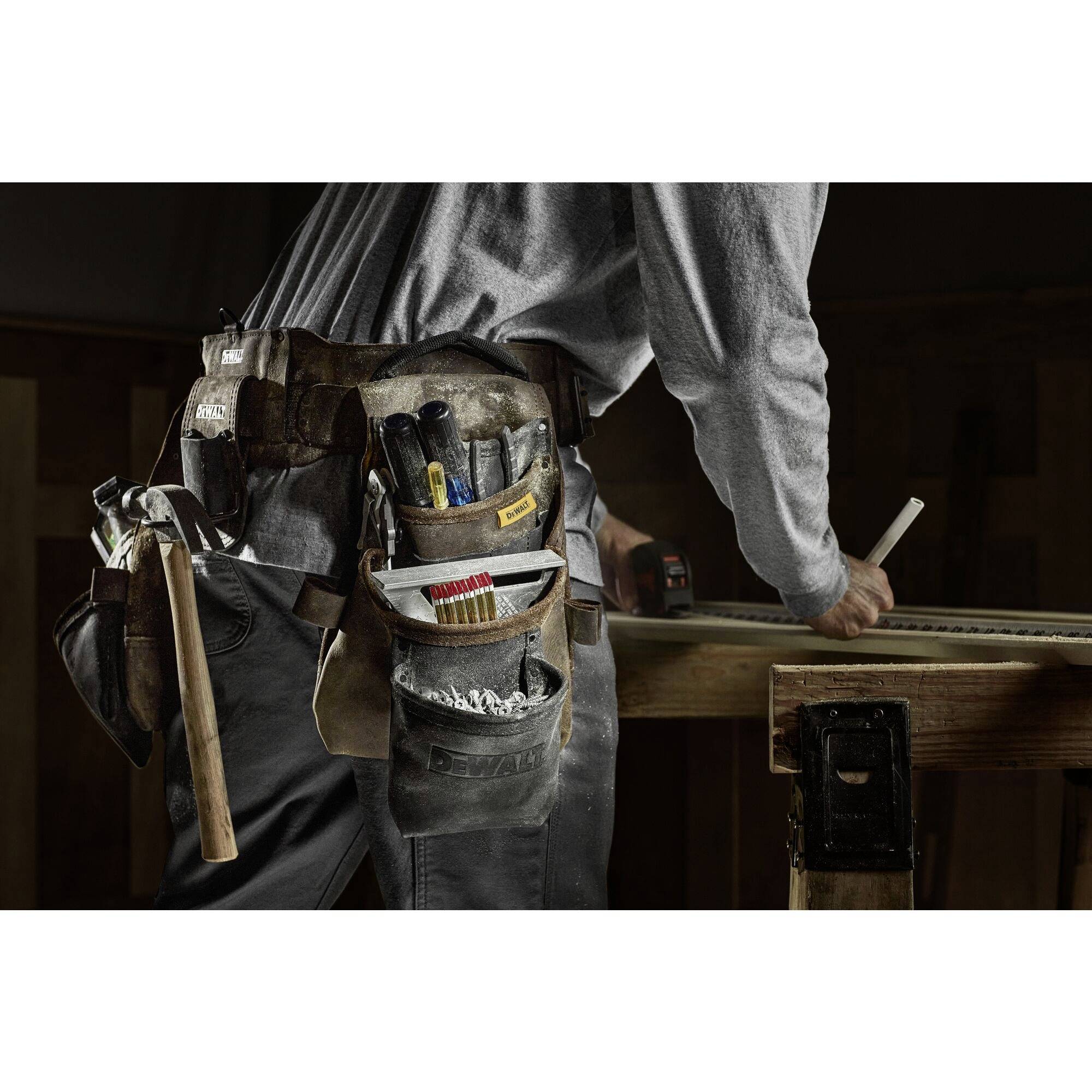 Osoba w stroju roboczym pracuje przy stole, mając na sobie pas narzędziowy. Widoczne są śrubokręty, szczypce i miara zwijana.