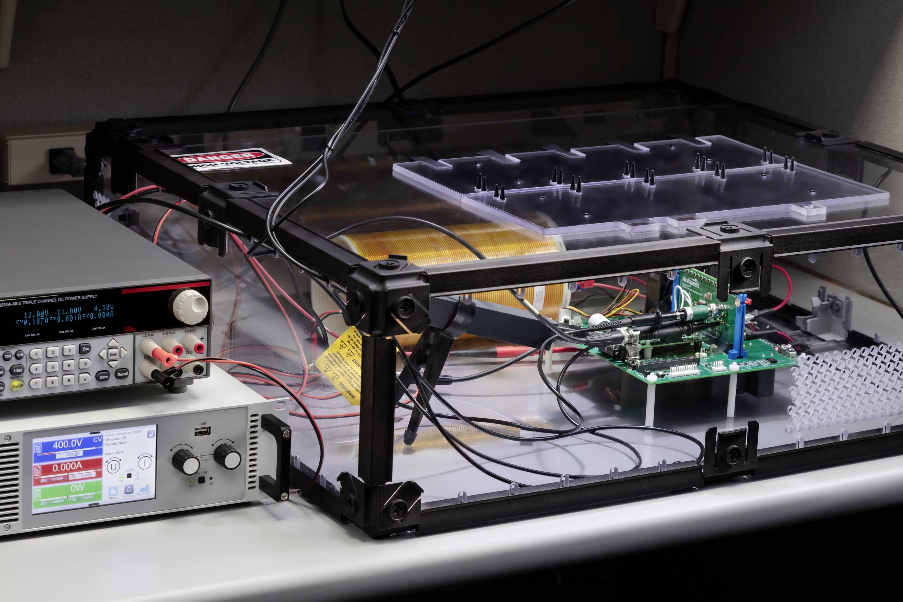 Układ testowy elektroniki na stole z urządzeniami i płytkami drukowanymi w przezroczystej obudowie, otoczony kablami i wyświetlaczami.