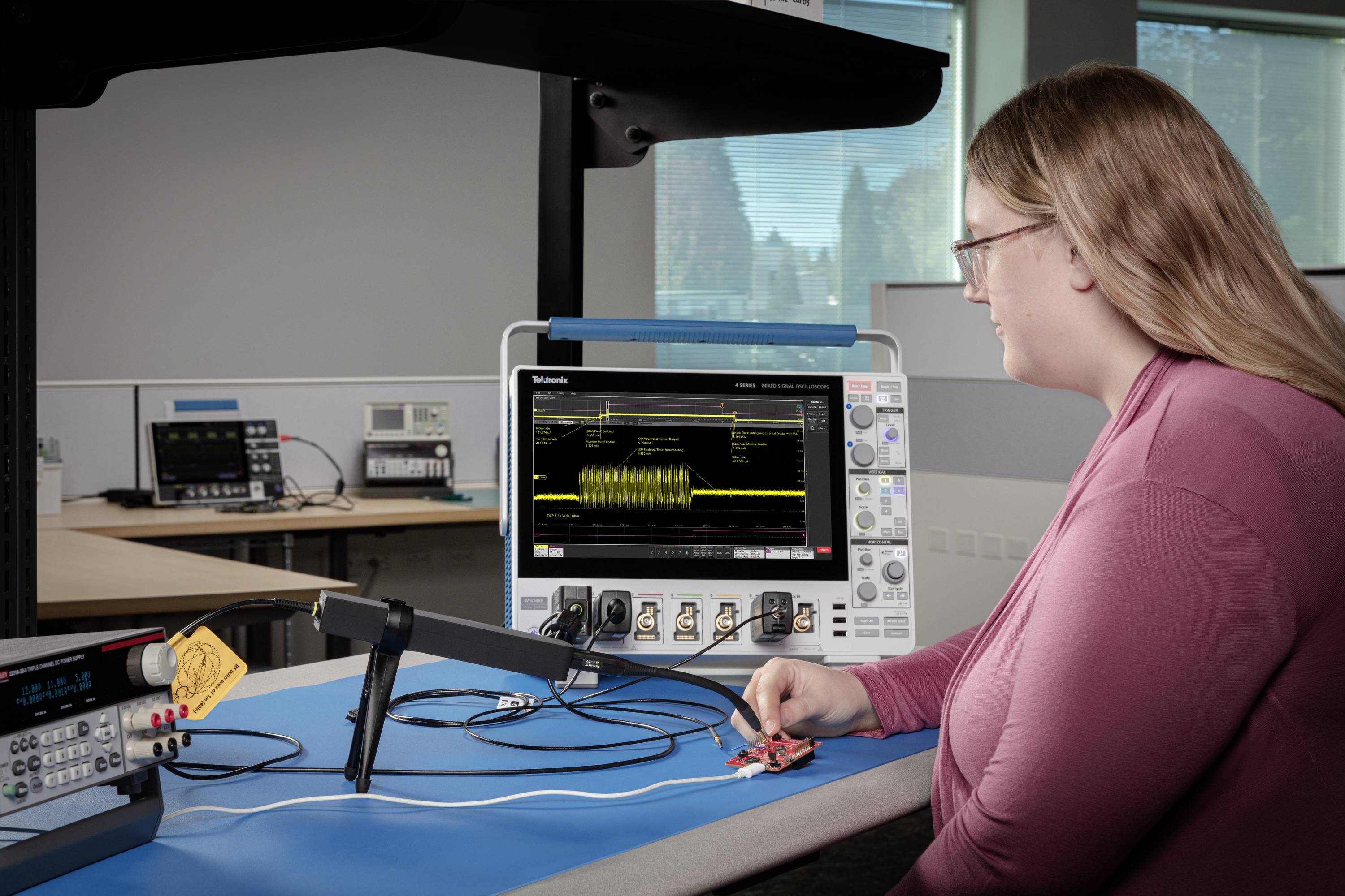 Osoba obsługuje oscyloskop w laboratorium. Urządzenie wyświetla na ekranie dane w postaci fal. W tle widoczne są kolejne urządzenia laboratoryjne.