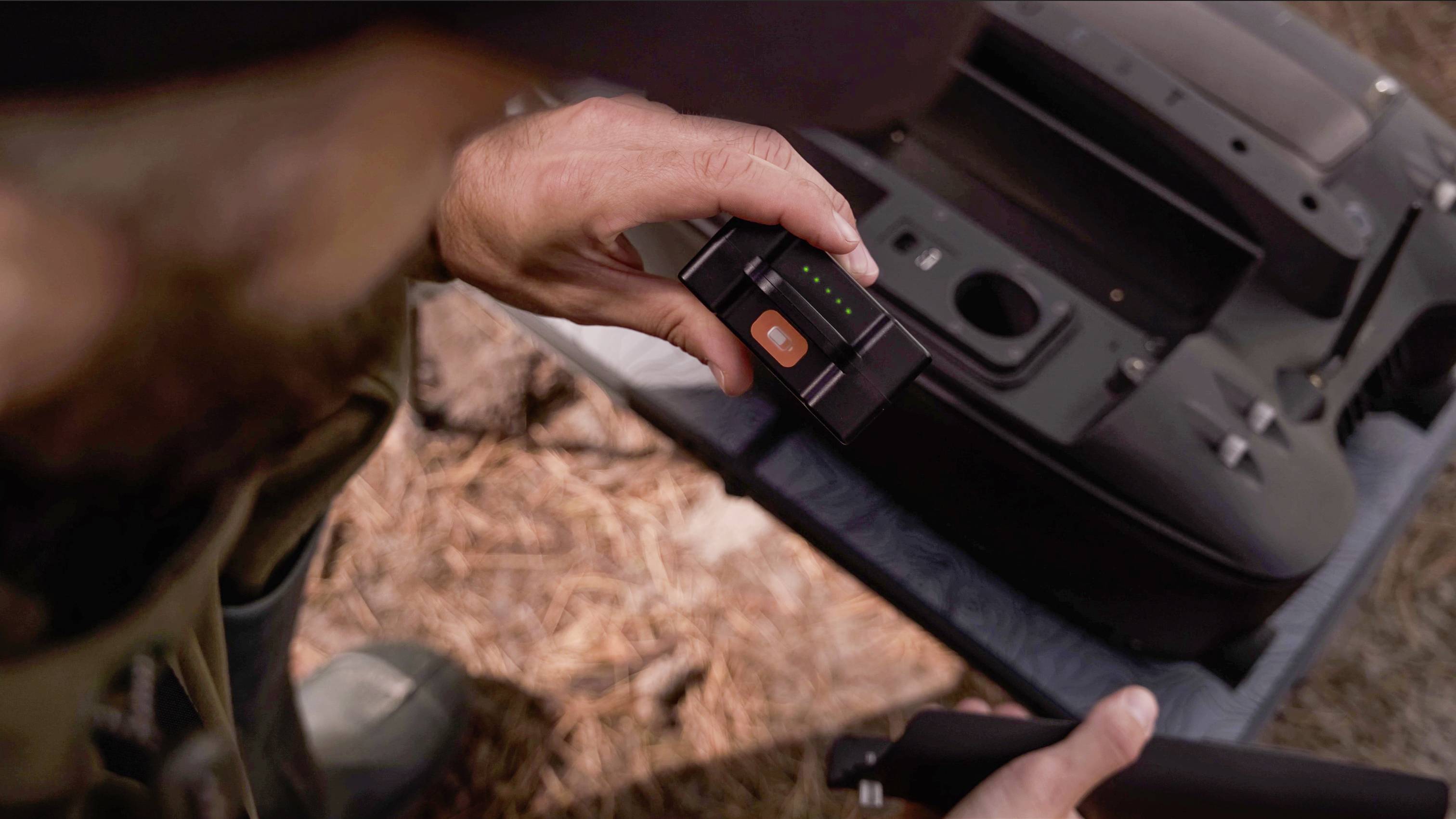 Osoba trzyma urządzenie elektroniczne z wyświetlaczem, na którym widoczny jest zielony pasek i pomarańczowy przycisk. W tle widać nieostre przedmioty.