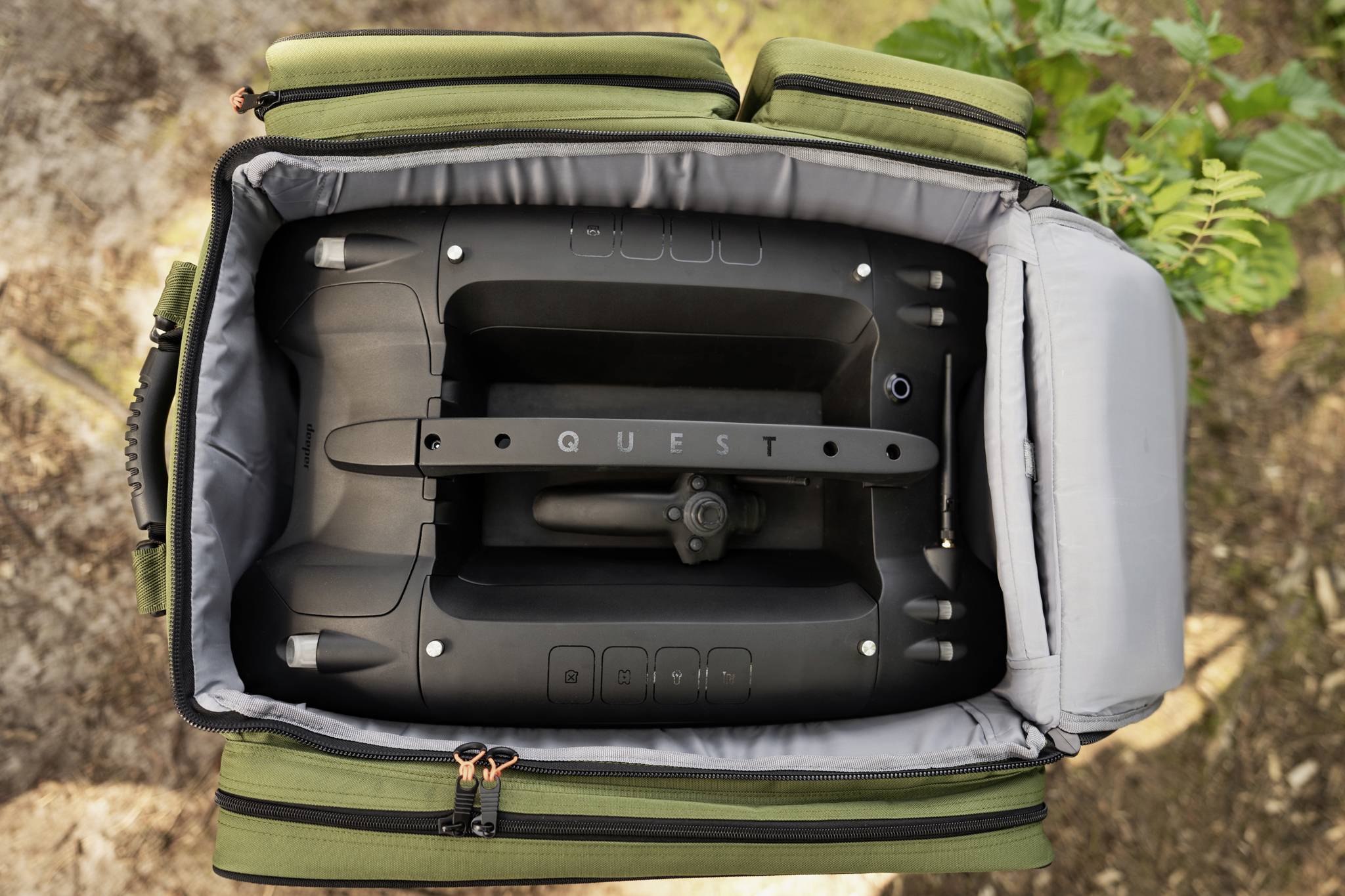 Czarne urządzenie z napisem 'QUEST' leży w otwartej, wyściełanej torbie. Na tle roślinnego otoczenia.