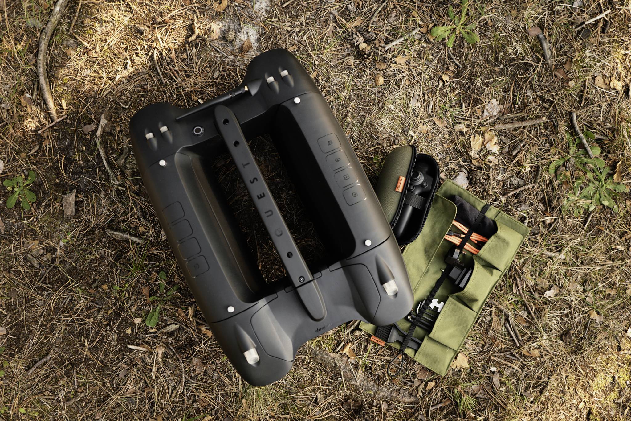 Czarny robot sprzątający leży na leśnej podłoże obok zielonej torby transportowej. Wydaje się być gotowy do użycia na wolnym powietrzu.