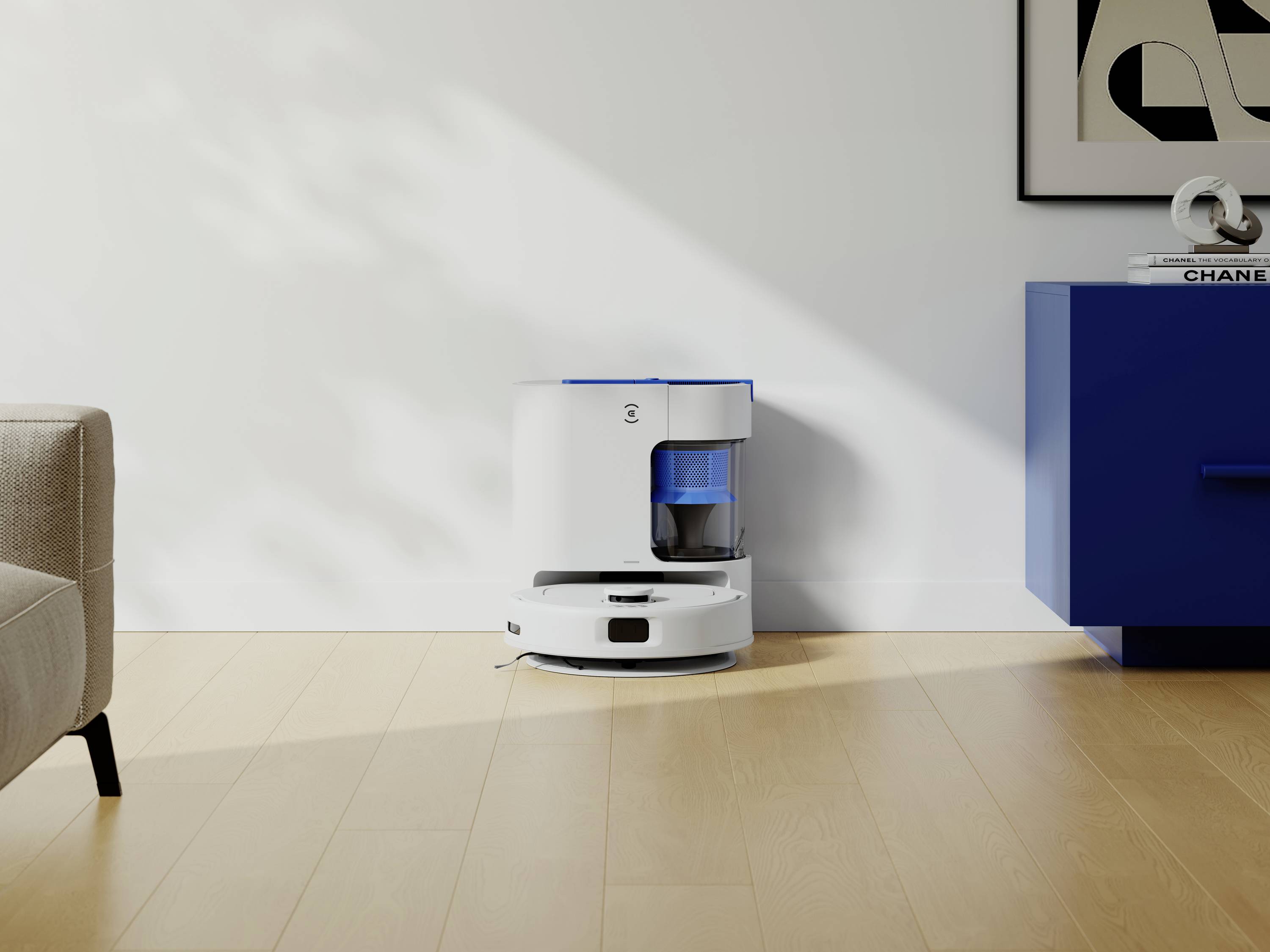 Biały robot sprzątający na drewnianej podłodze, obok niebieskiej komody i sofy. Na ścianie wisi abstrakcyjne dzieło sztuki.