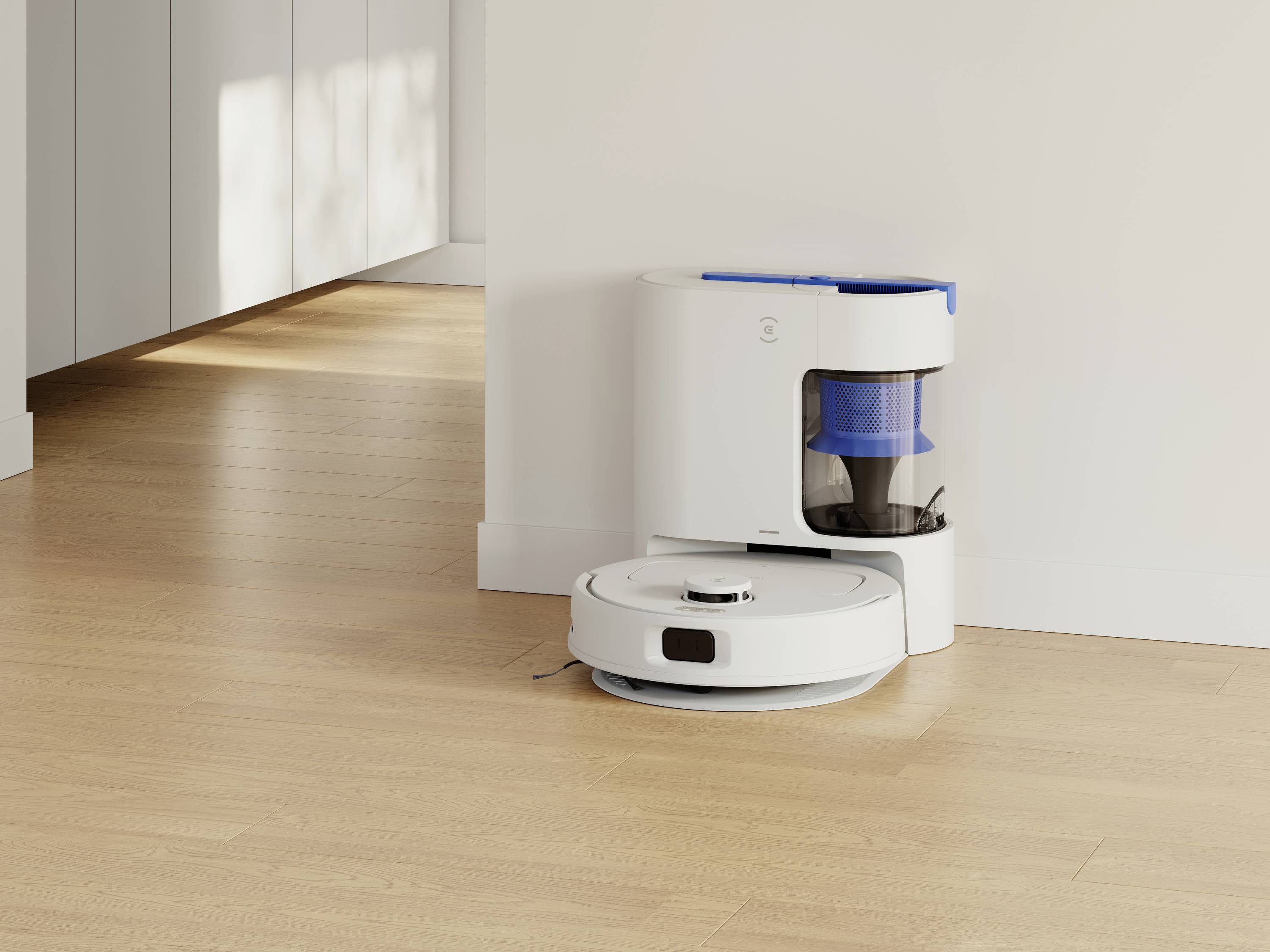 Biały robot odkurzający cumuje przy stacji ładującej w jasnym pomieszczeniu z drewnianą podłogą. Brak osób w kadrze.