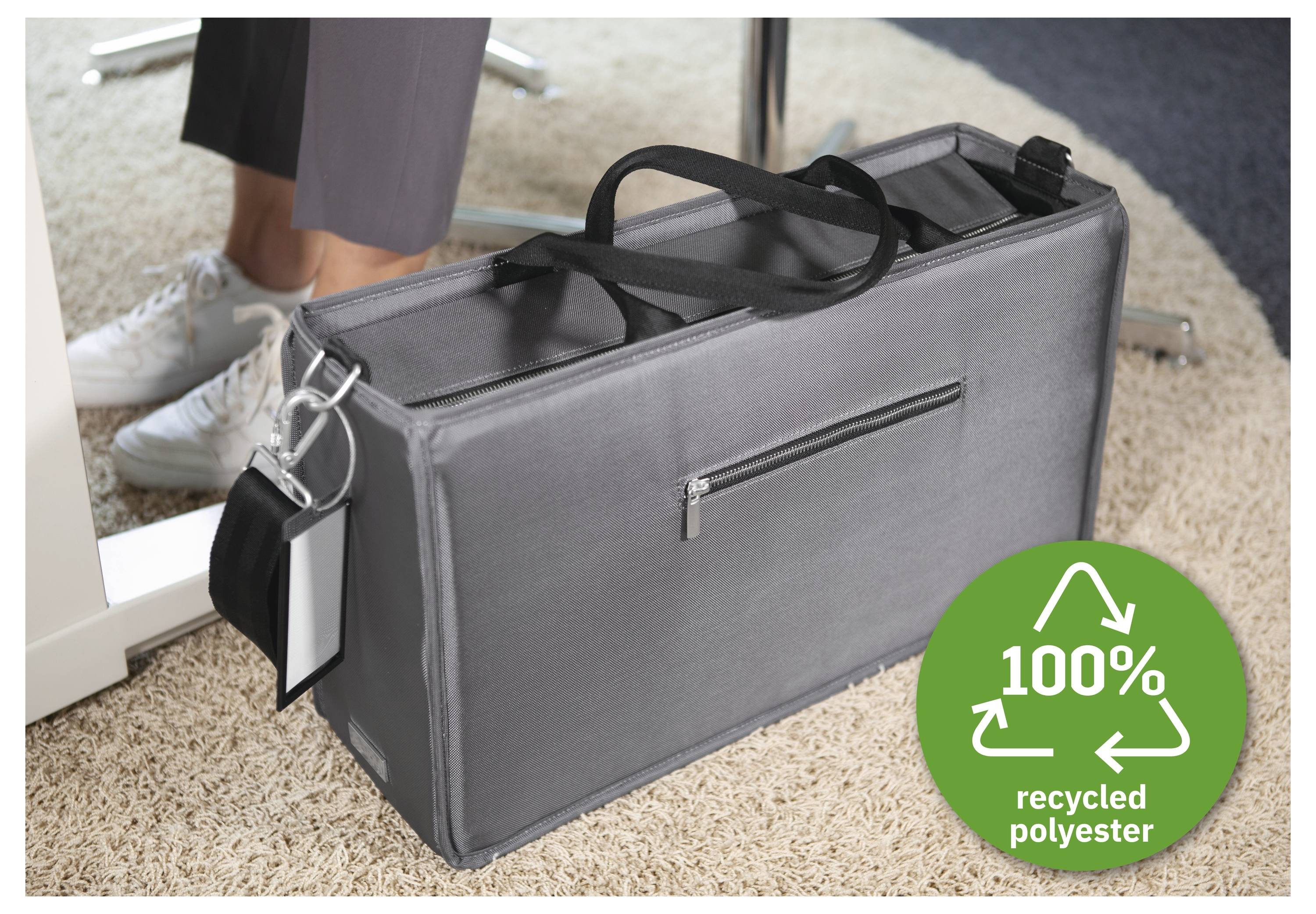 Szara torba podróżna wykonana z 100% recyklingowanego poliestru, pokazana z paskiem do noszenia i kieszenią na zamek błyskawiczny na dywanie.