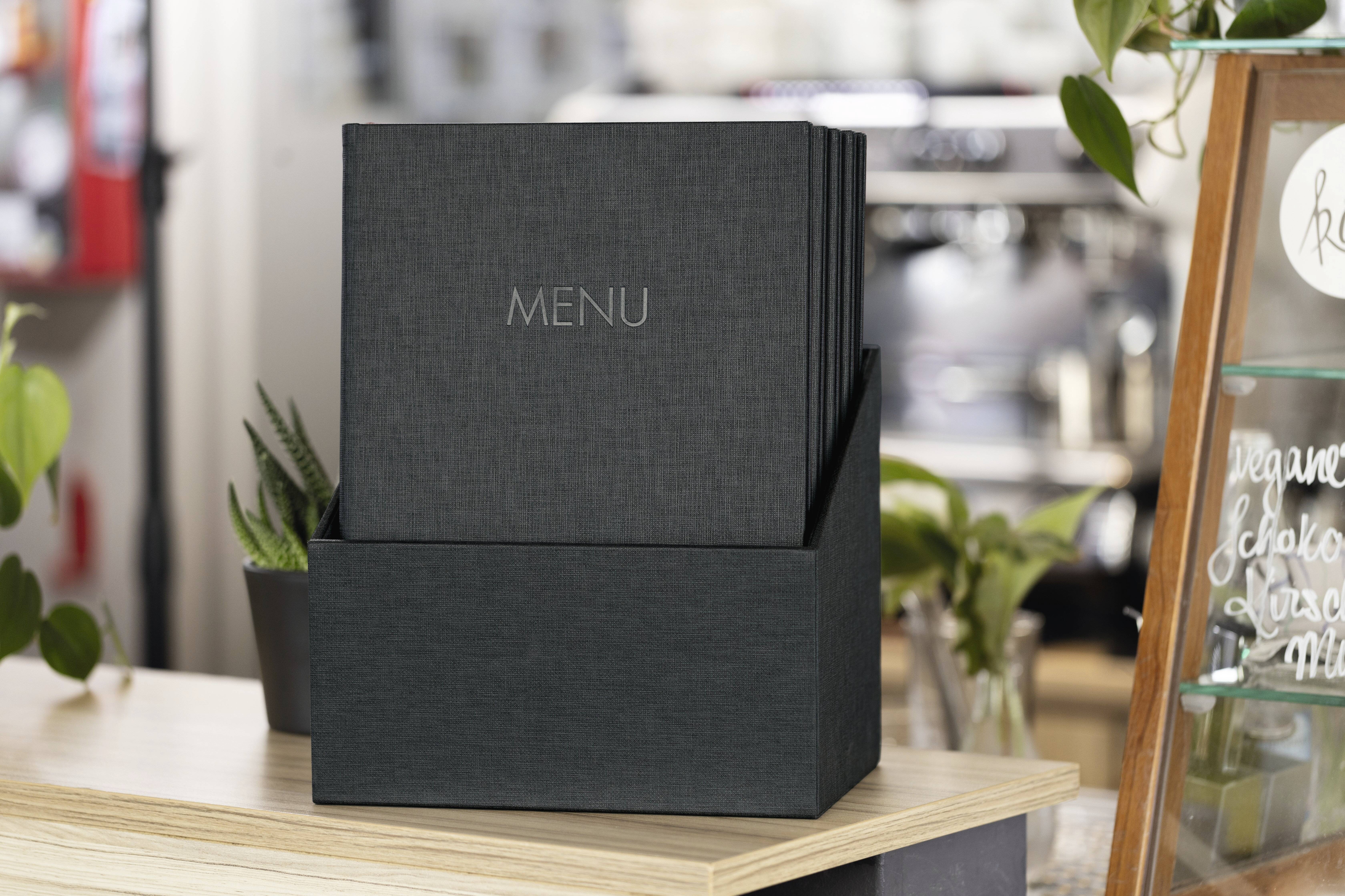 Czarny materiałowy stojak na menu stoi na stole, otoczony roślinami, w tle rozmyte wnętrze kawiarni.