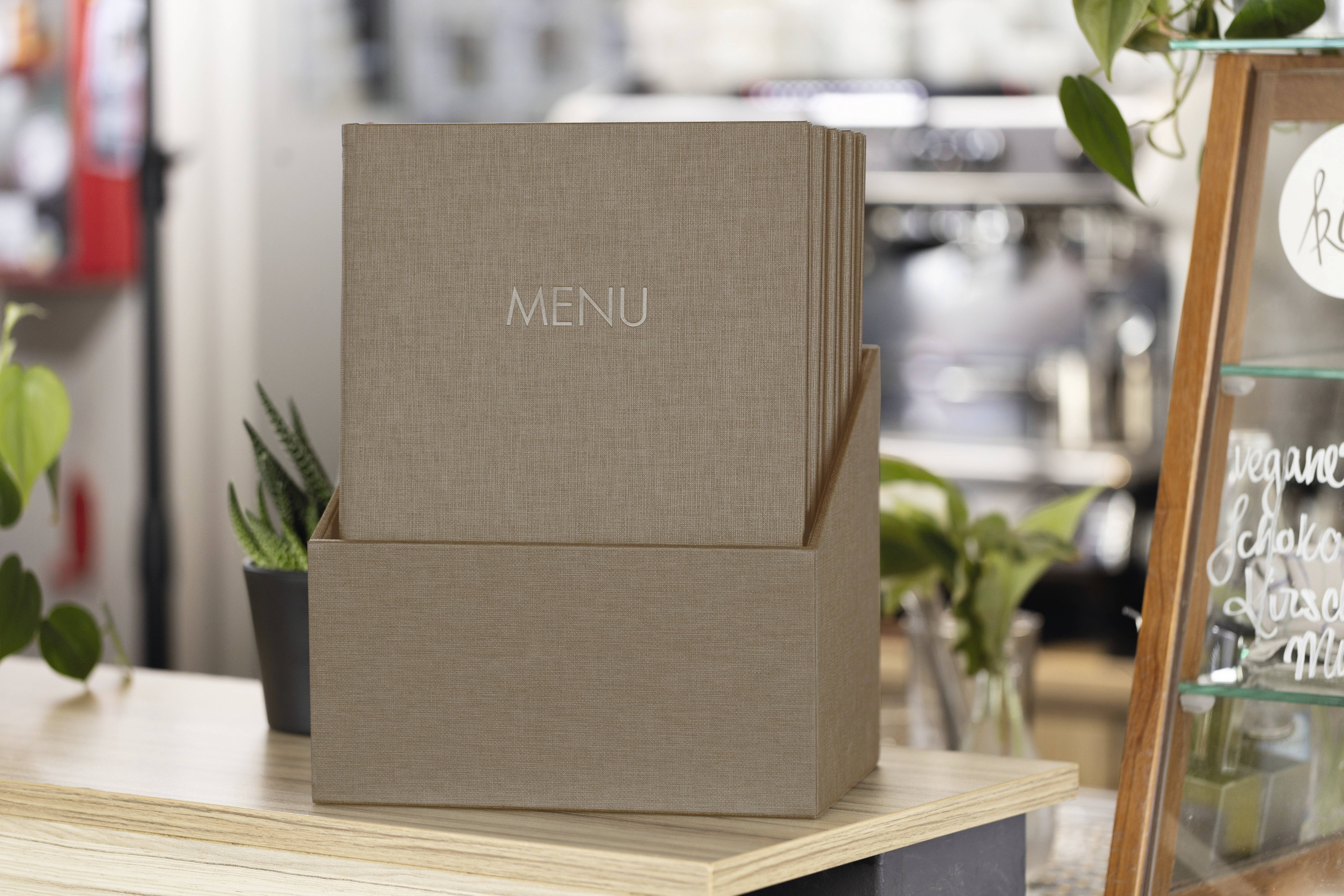 Okładki menu w etui na karty na stoliku w kawiarni; w tle widoczne są rośliny i rozmyte wyposażenie kawiarni.