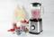 Blender zawiera czerwony smoothie owocowy. Obok stoją napełnione szklanki, świeże truskawki, jagody oraz łyżka.