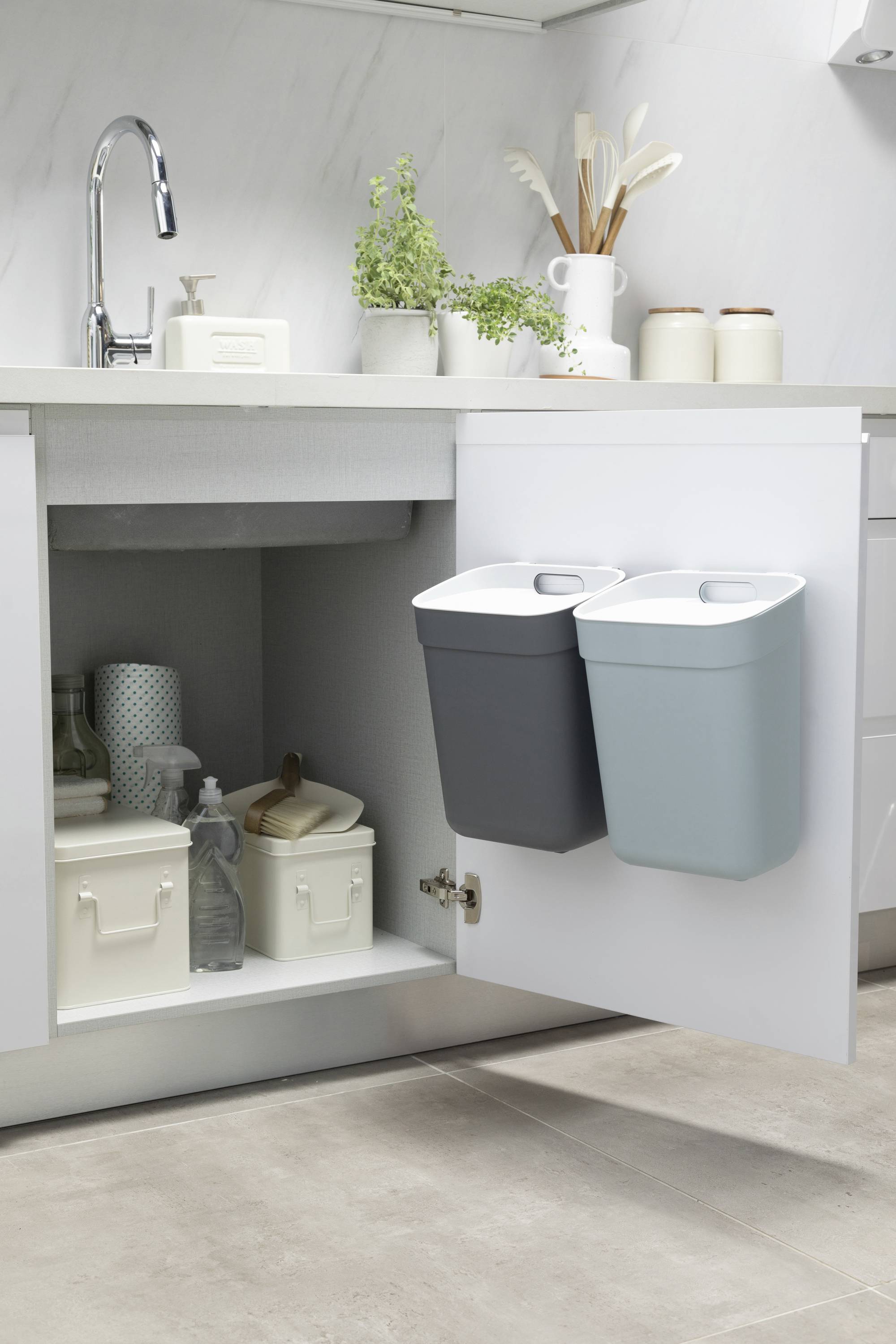 Szafka pod zlewem z otwartymi drzwiczkami prezentuje dwie wiszące kosze na śmieci oraz pudełka do przechowywania. Starannie zorganizowana kuchnia.