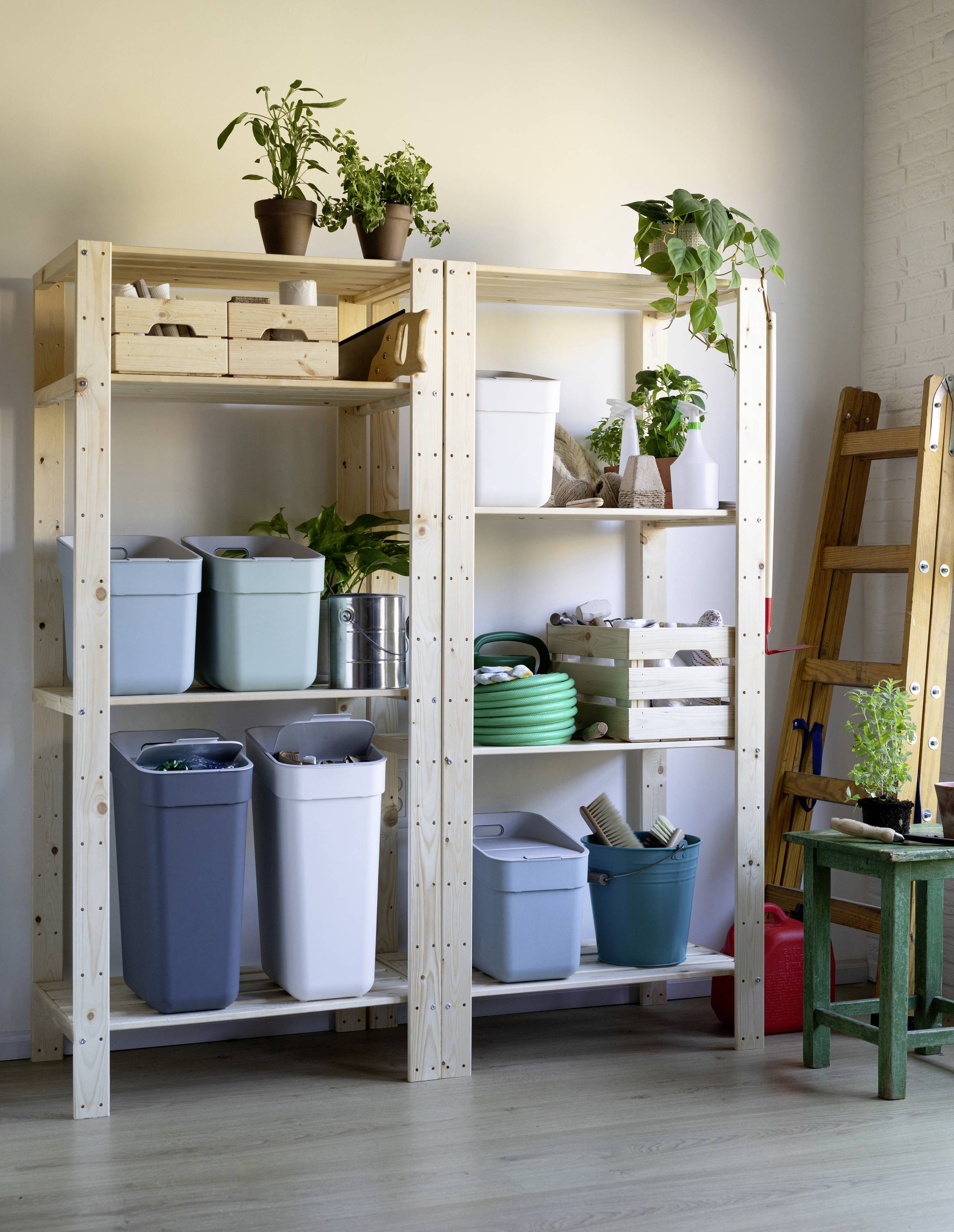 Drewniany regał z roślinami na górnych półkach i kolorowe kosze na śmieci poniżej. Obok stoi drabina.