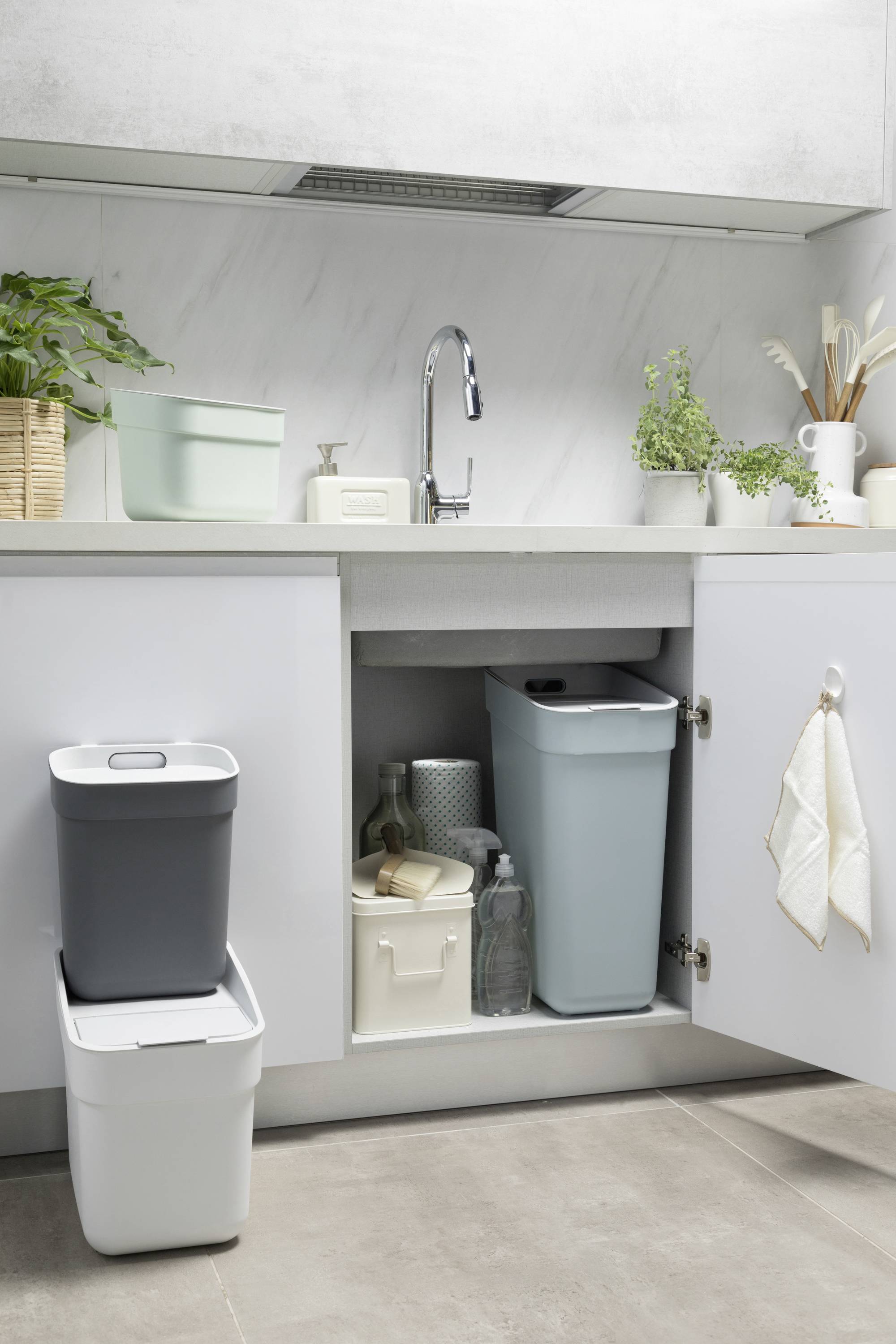 Pod zlewem kuchennym stoją kosze na śmieci i pojemniki na zapasy. Na blacie kuchennym znajdują się rośliny i inne przedmioty gospodarstwa domowego.