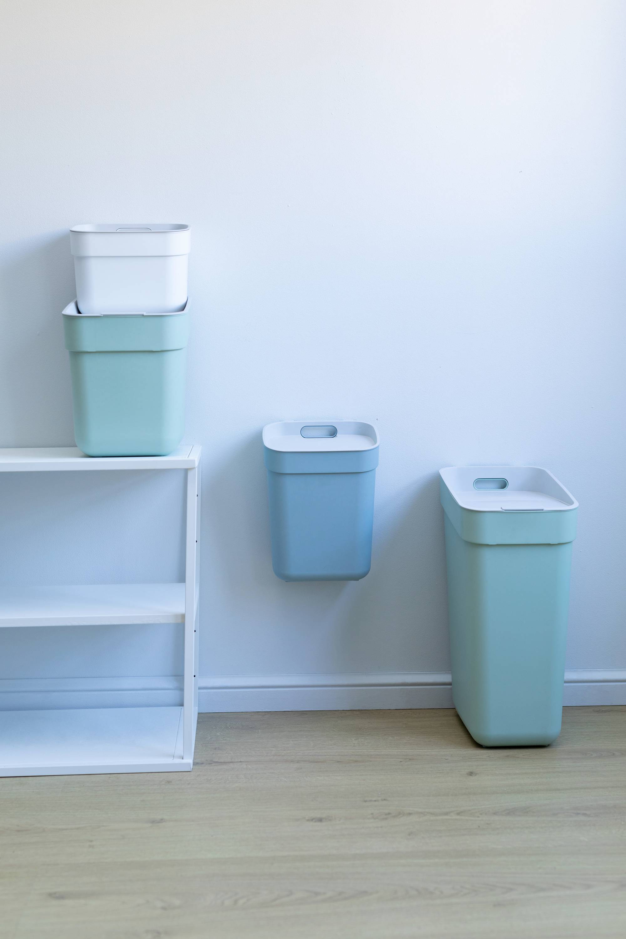 Pojemniki na odpady segregowane w różnych rozmiarach przed białą ścianą; dwa pojemniki ułożone jeden na drugim, jeden zamocowany na ścianie, jeden obok.