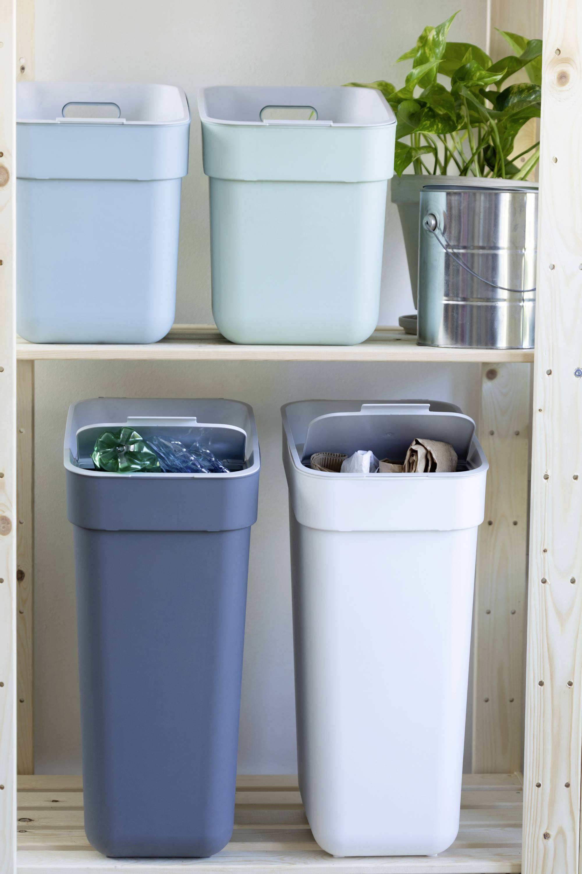 Cztery kosze na śmieci w różnych kolorach stoją na półce. Na górze znajdują się lekkie pojemniki, na dole są wypełnione materiałami do recyklingu. Obok stoi mała zielona roślina.