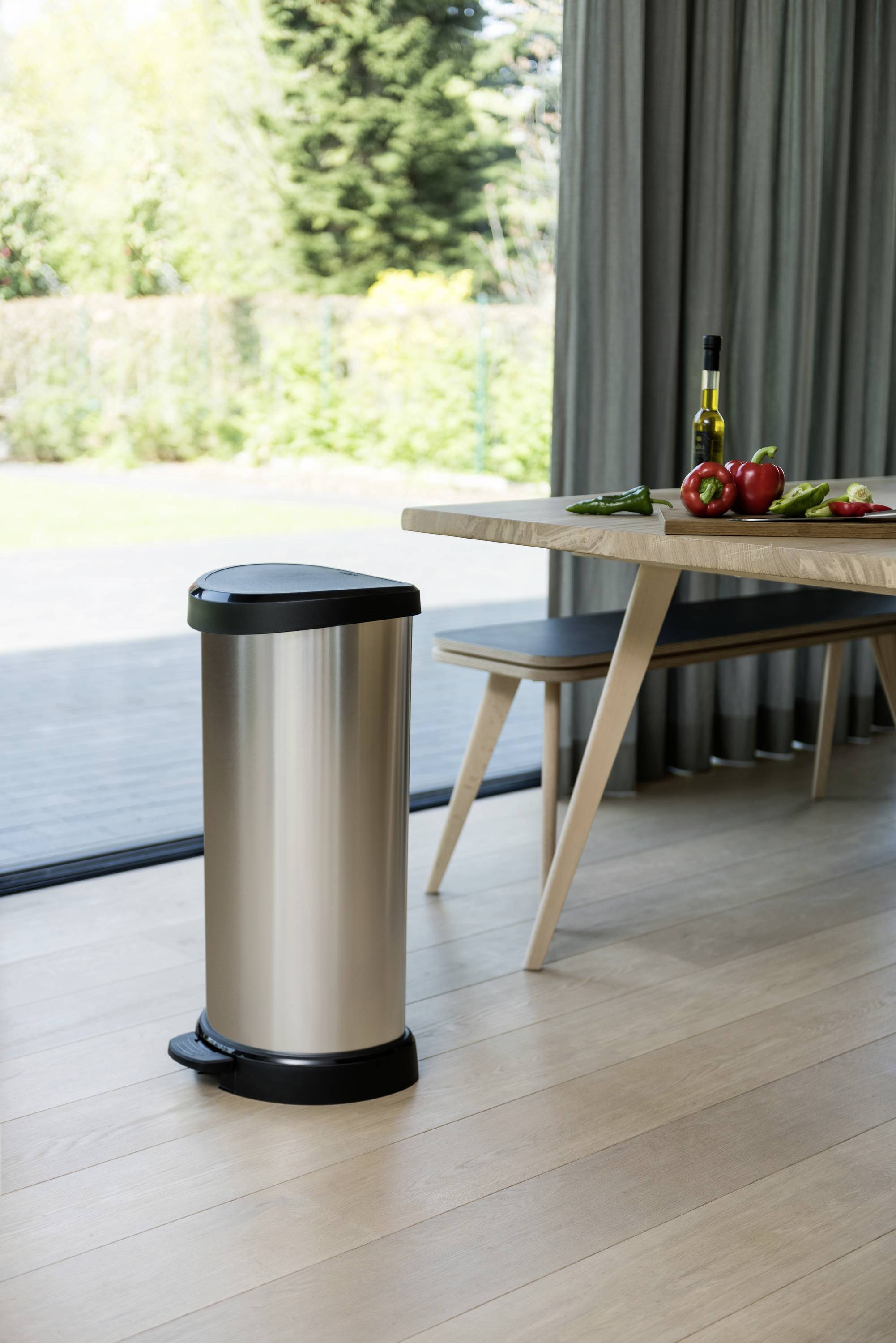 Nowoczesny kosz na śmieci stoi w jasnej kuchni obok stołu, na którym leżą warzywa i butelka wina.