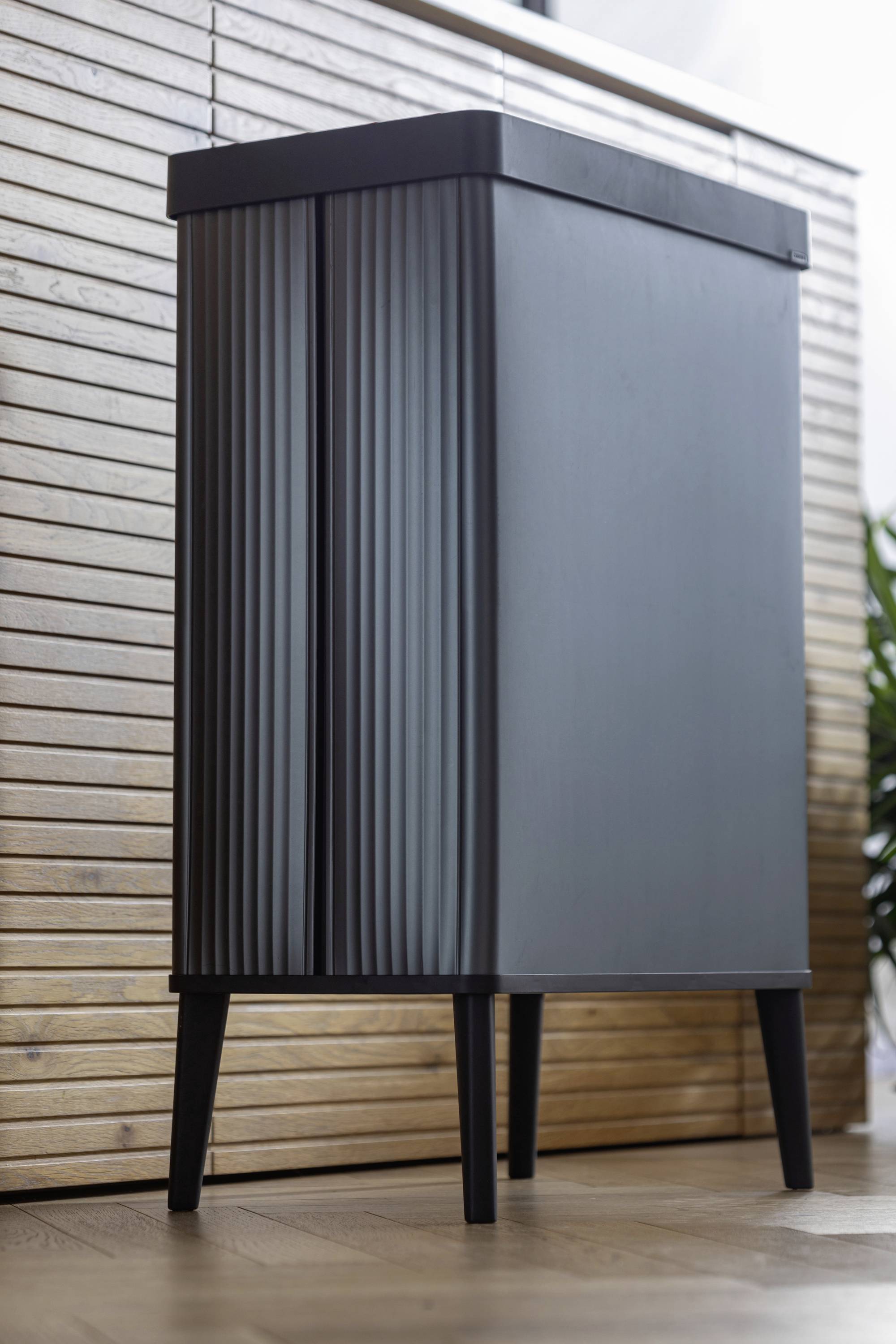 Nowoczesna, wysoka komoda o ciemnej, żłobionej powierzchni, stojąca na czterech nogach przed drewnianą ścianą.