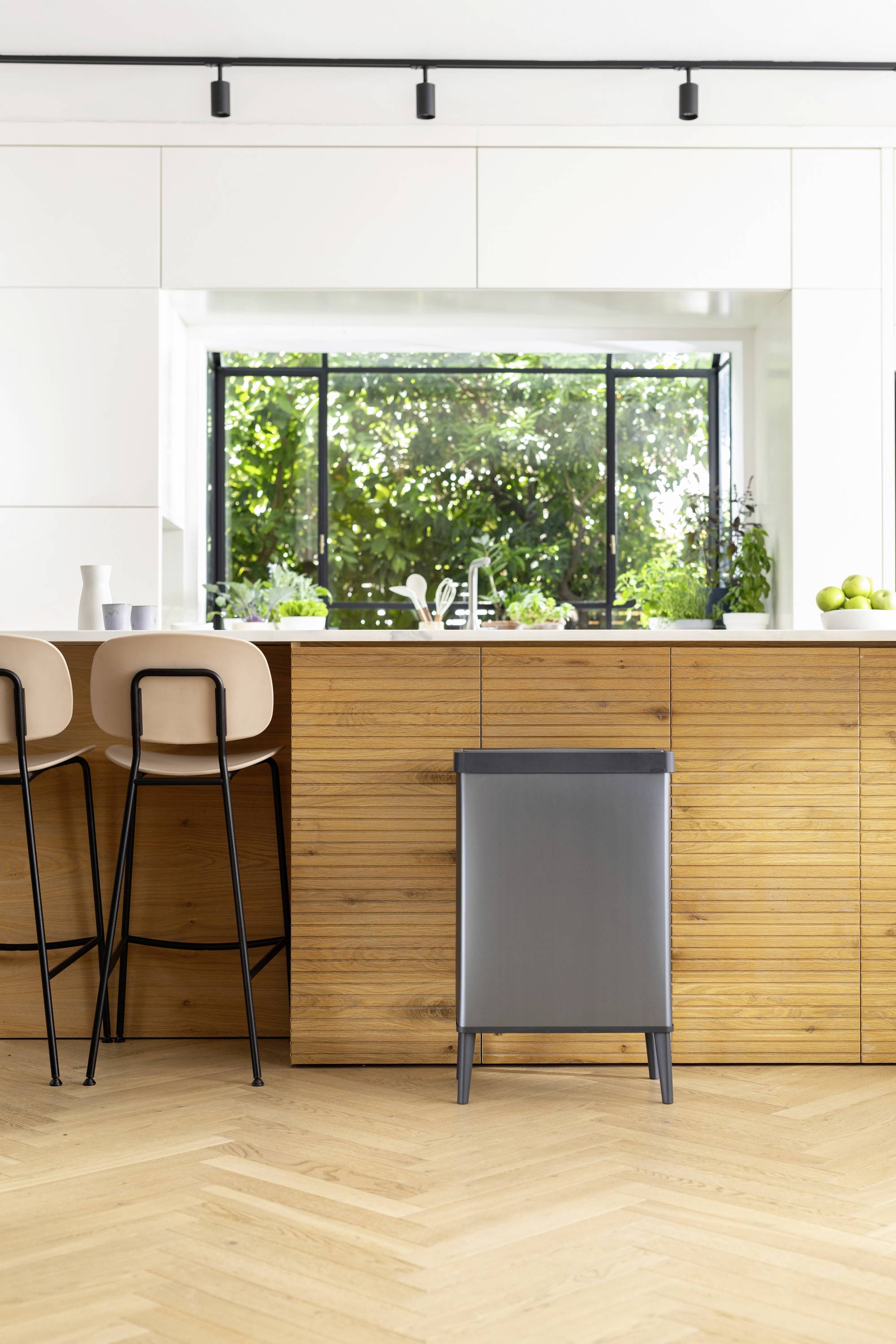 Nowoczesna kuchnia z drewnianą wyspą, dwoma stołkami barkowymi i dużym oknem, przez które wpada obfite światło dzienne. Na zewnątrz widoczne są zielone rośliny.