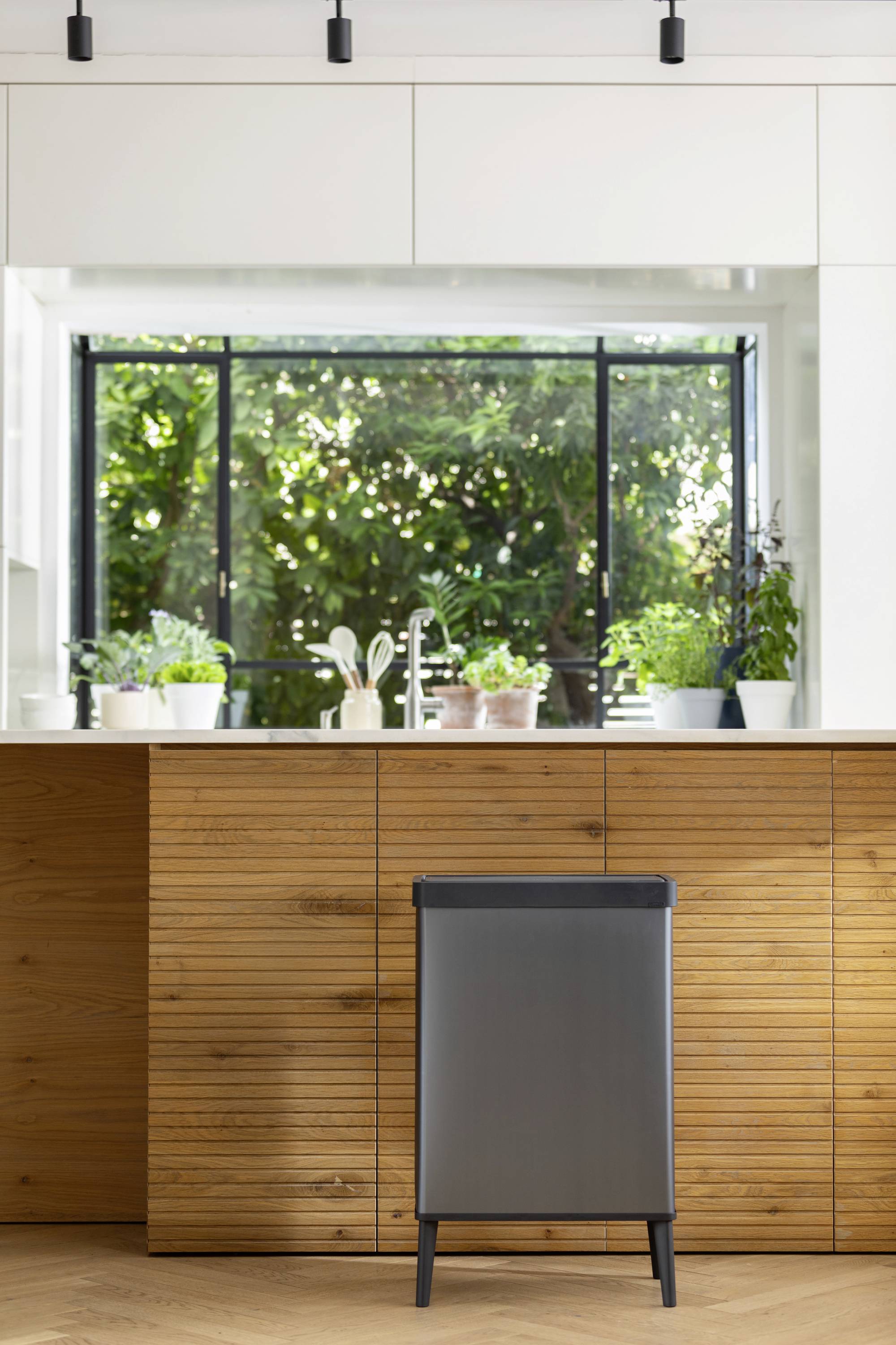 Nowoczesna kuchnia z drewnianą zabudową i dużym oknem. Na blacie stoją doniczki z roślinami, a w przedniej części widoczny jest kosz na śmieci.
