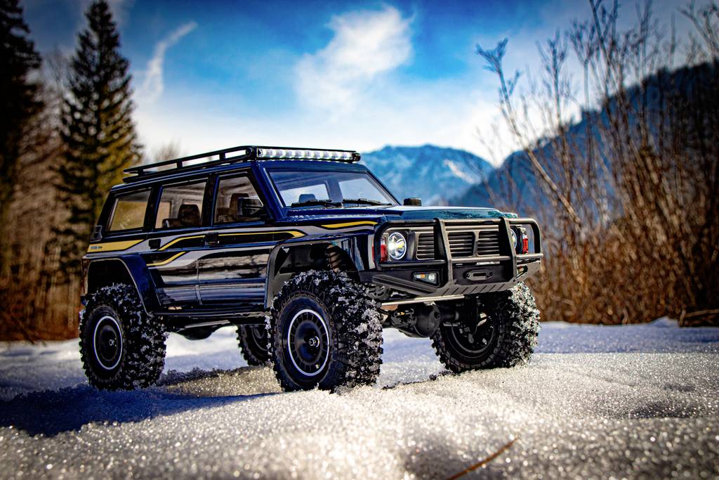 Czarny zdalnie sterowany samochód terenowy jedzie po zaśnieżonym terenie na tle zalesionych gór i błękitnego nieba.