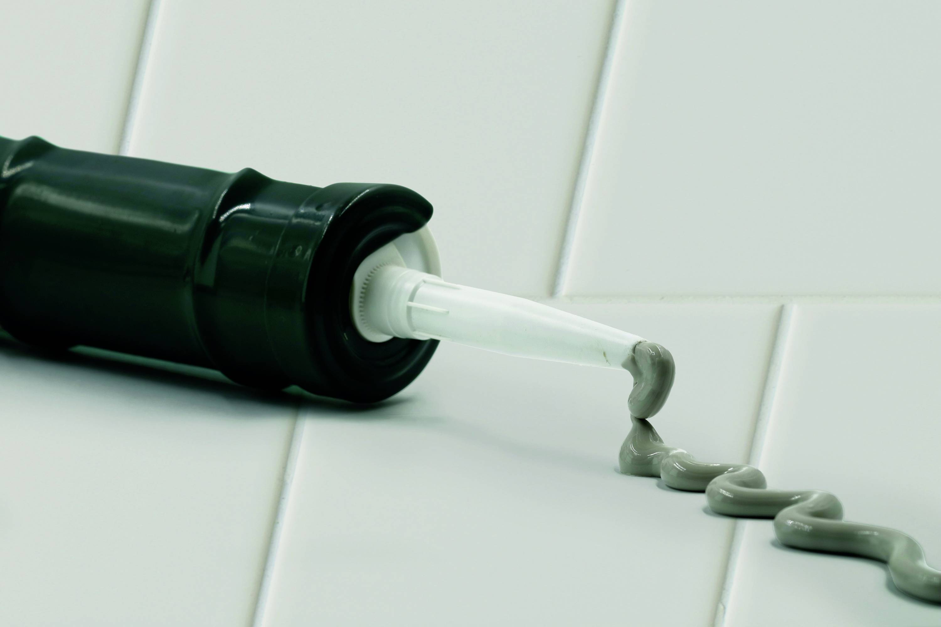 Czarna pistolet do kartusza leży na białych kafelkach i nakłada szarą masę uszczelniającą w kształcie linii S.