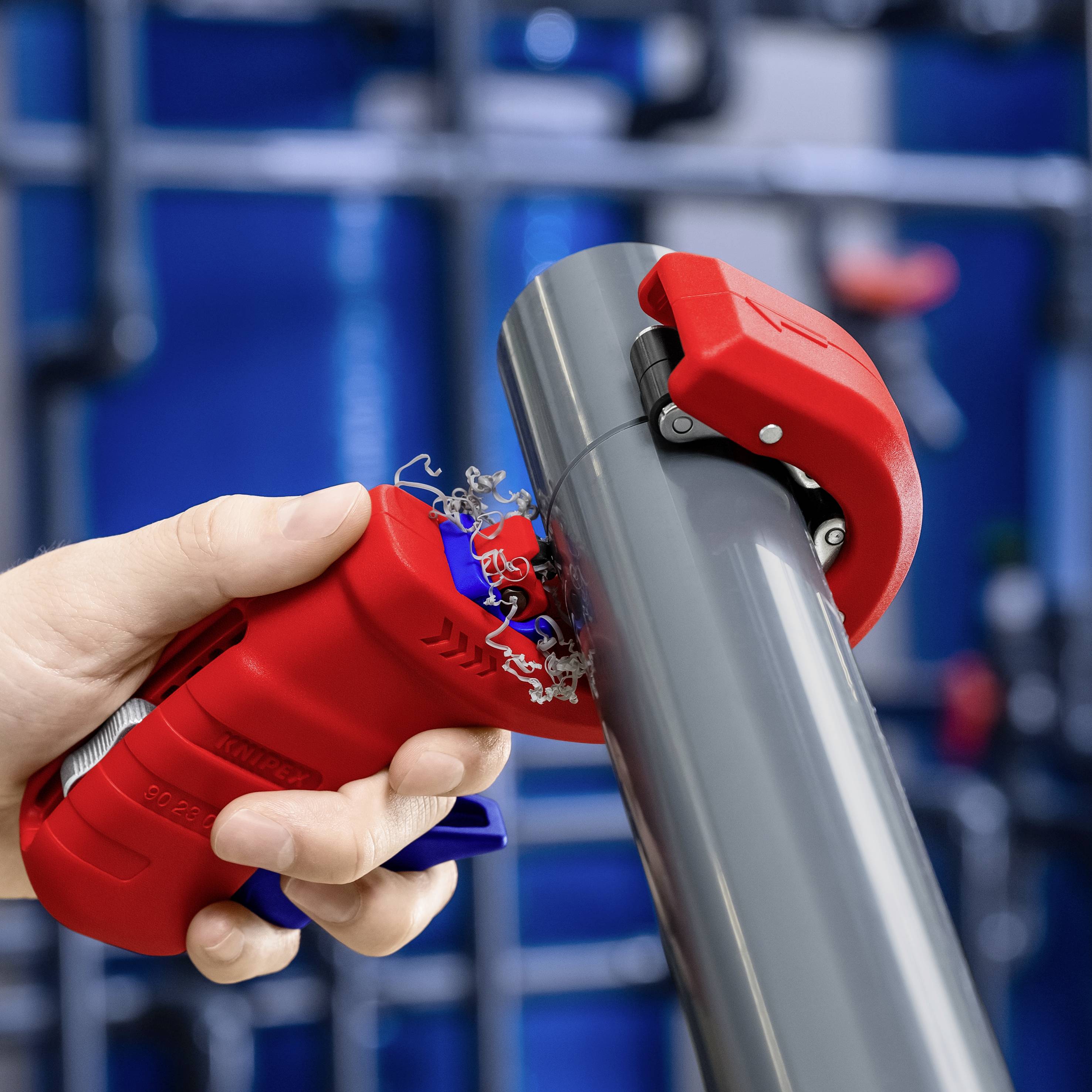 Osoba ucina szare rury za pomocą czerwonego obcinaka do rur. W tle nieostre, przemysłowe rurociągi.