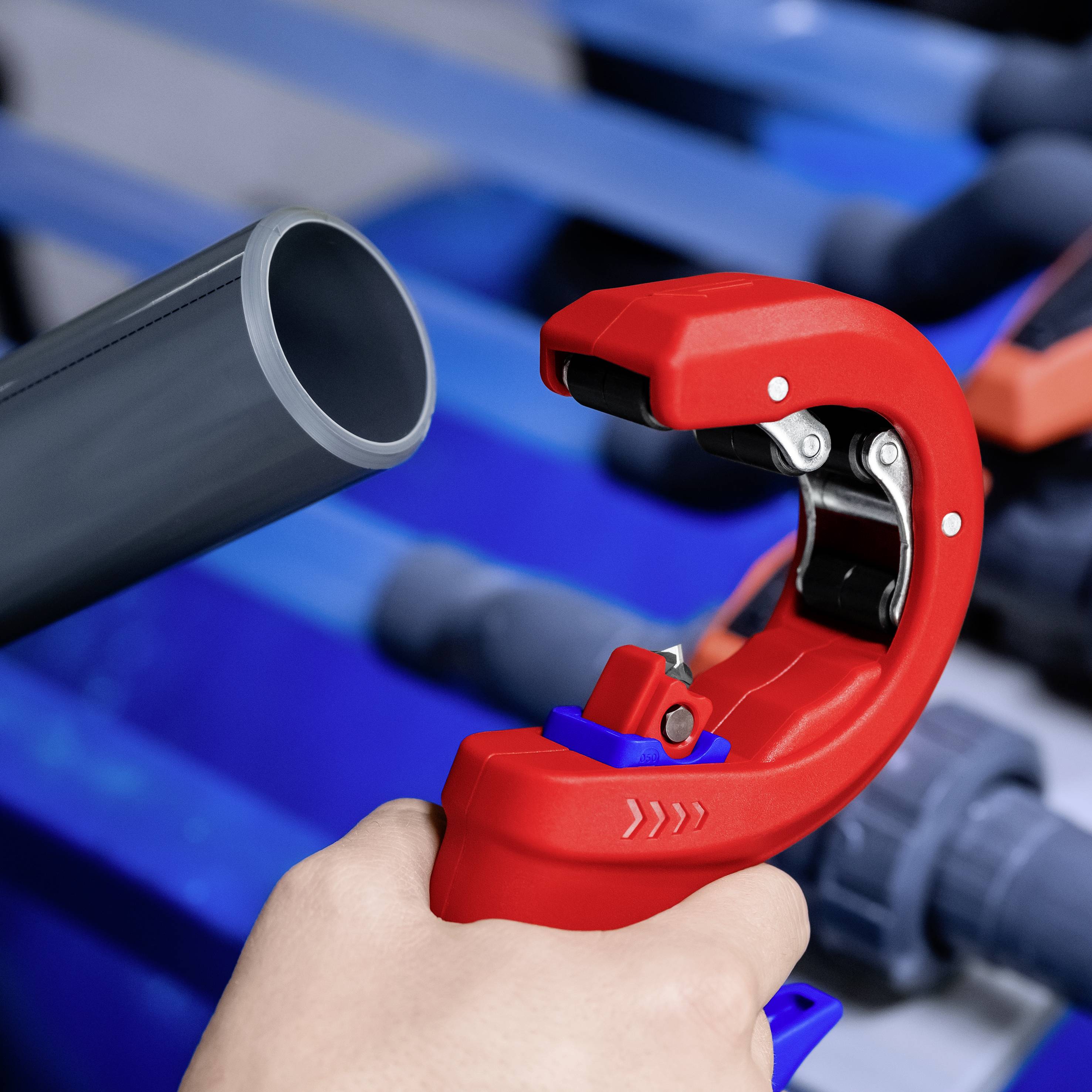 Czerwone nożyce do rur przecinają szare rury z tworzywa sztucznego. Tło jest rozmyte, dominują odcienie niebieskiego i szarego. Ręka robotnika trzyma rurę.