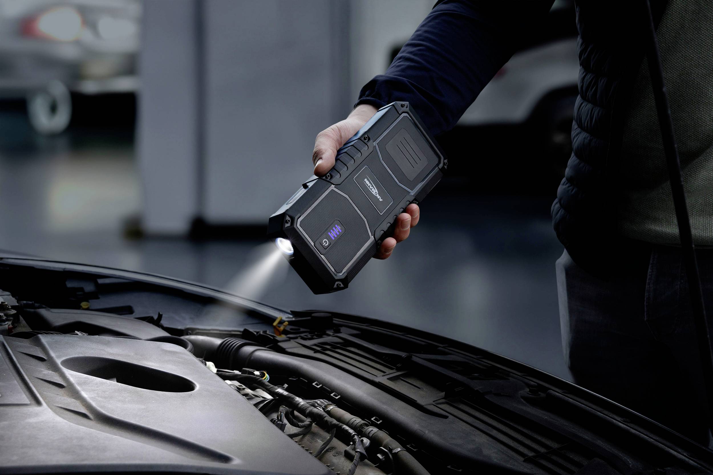 Osoba trzyma przenośną lampę roboczą nad otwartą komorą silnika w warsztacie. Lampa oświetla obszar silnika.