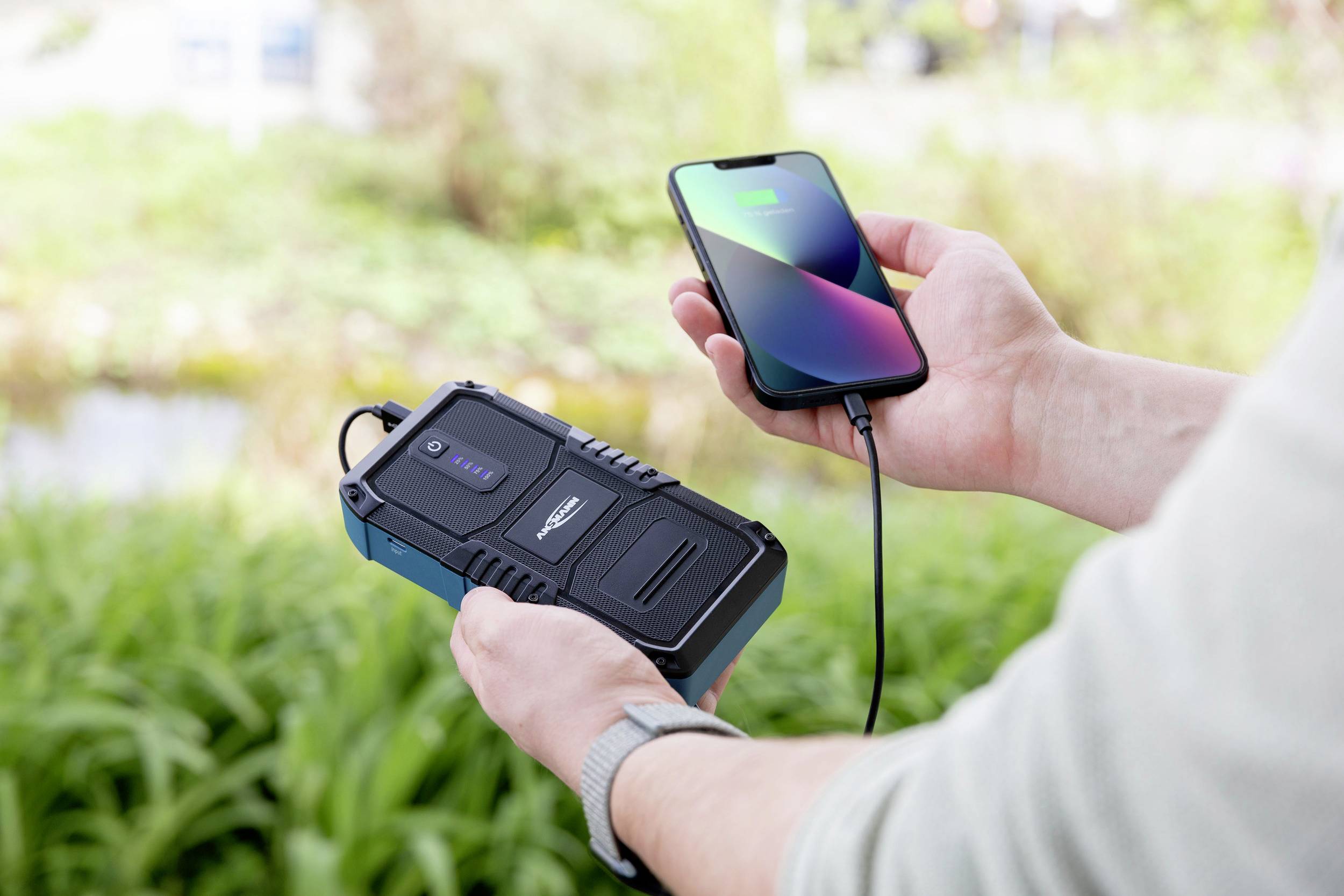 Osoba ładuje smartfon na zewnątrz za pomocą przenośnego powerbanku; powerbank wskazuje pełne naładowanie.