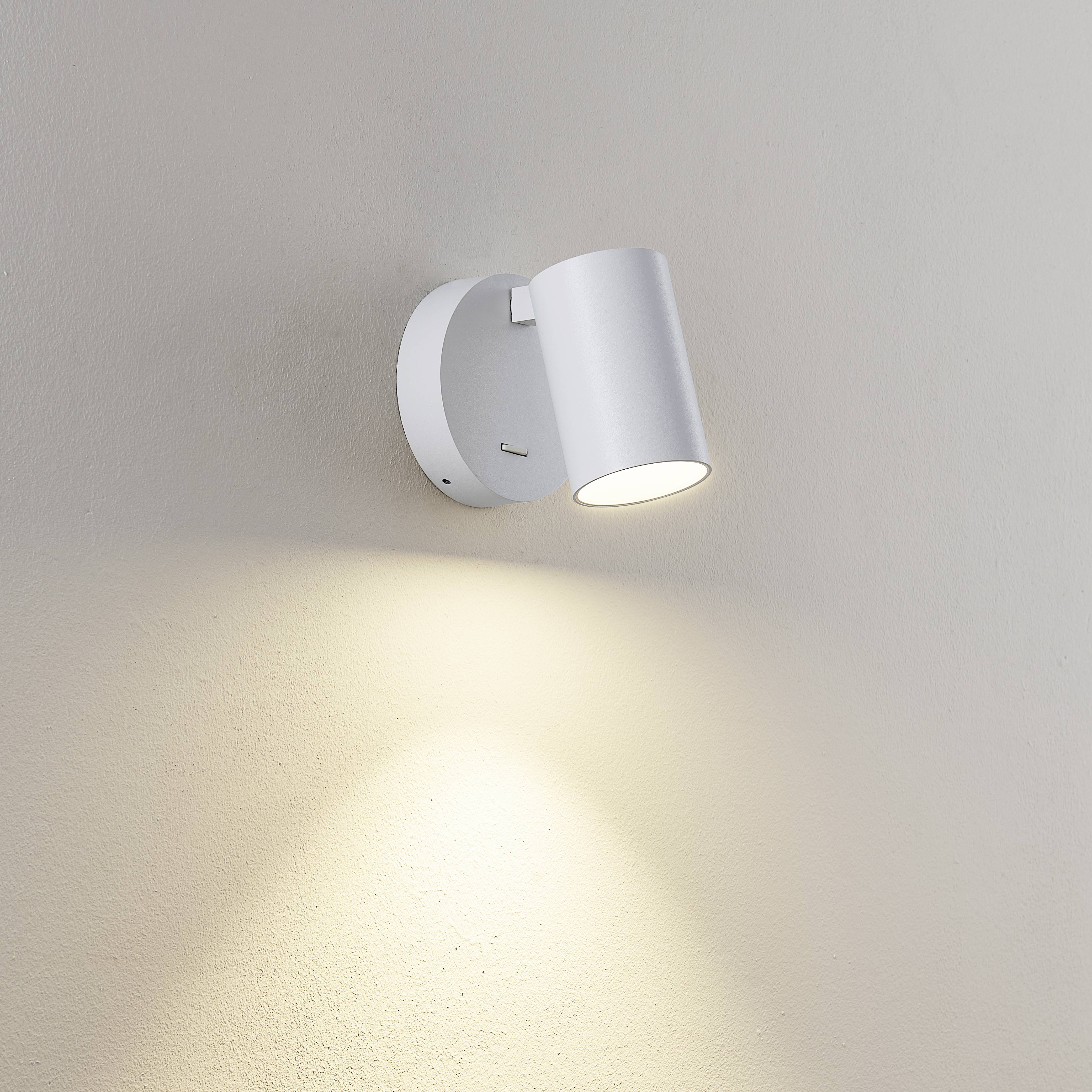 Kinkiet o cylindrycznym kształcie wykonany z białego metalu, świecący w dół i delikatnie oświetlający beżową ścianę.