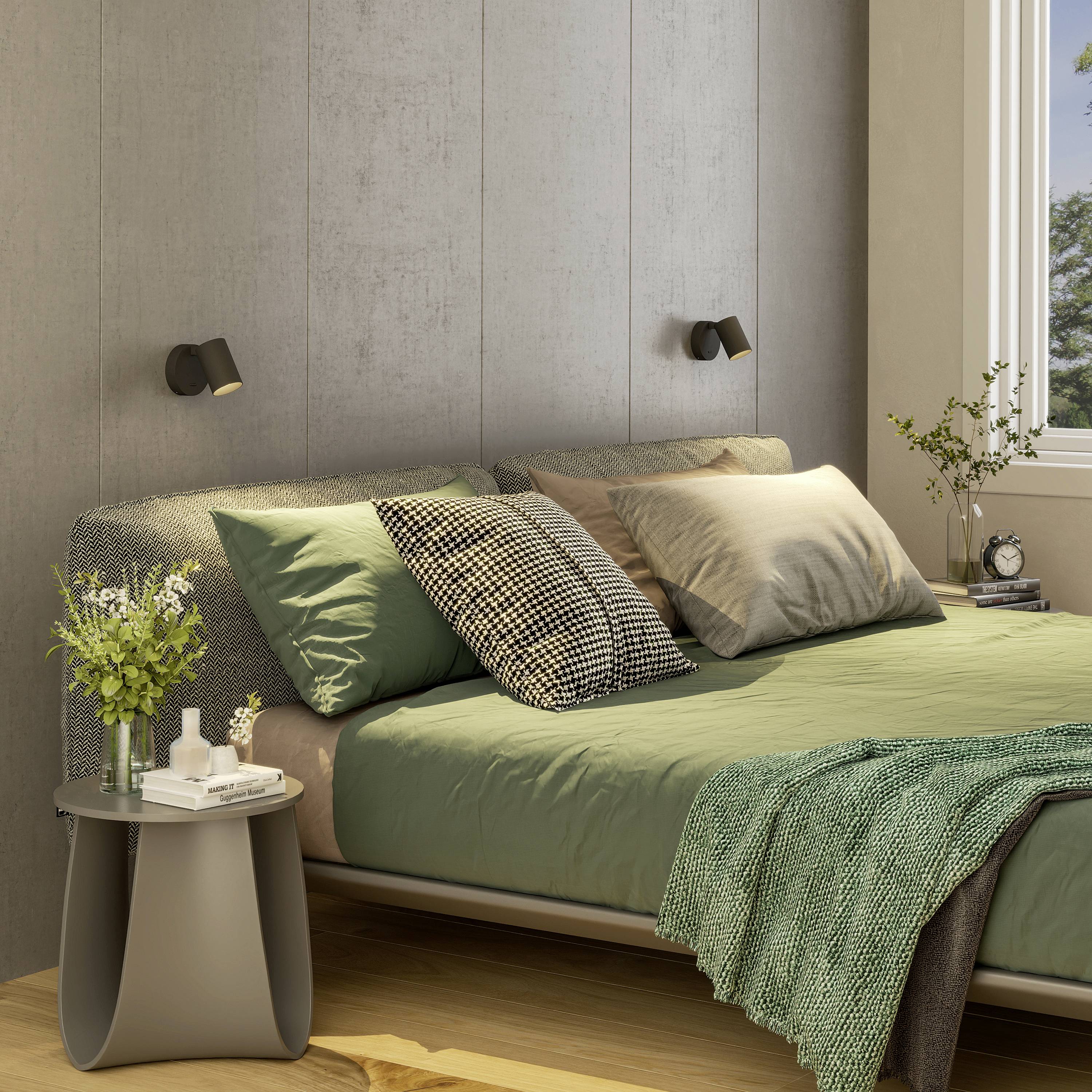 Przytulna sypialnia z podwójnym łóżkiem, szarymi i zielonymi narzutami, poduszkami, roślinami na stolikach nocnych oraz nowoczesnym oświetleniem ściennym.