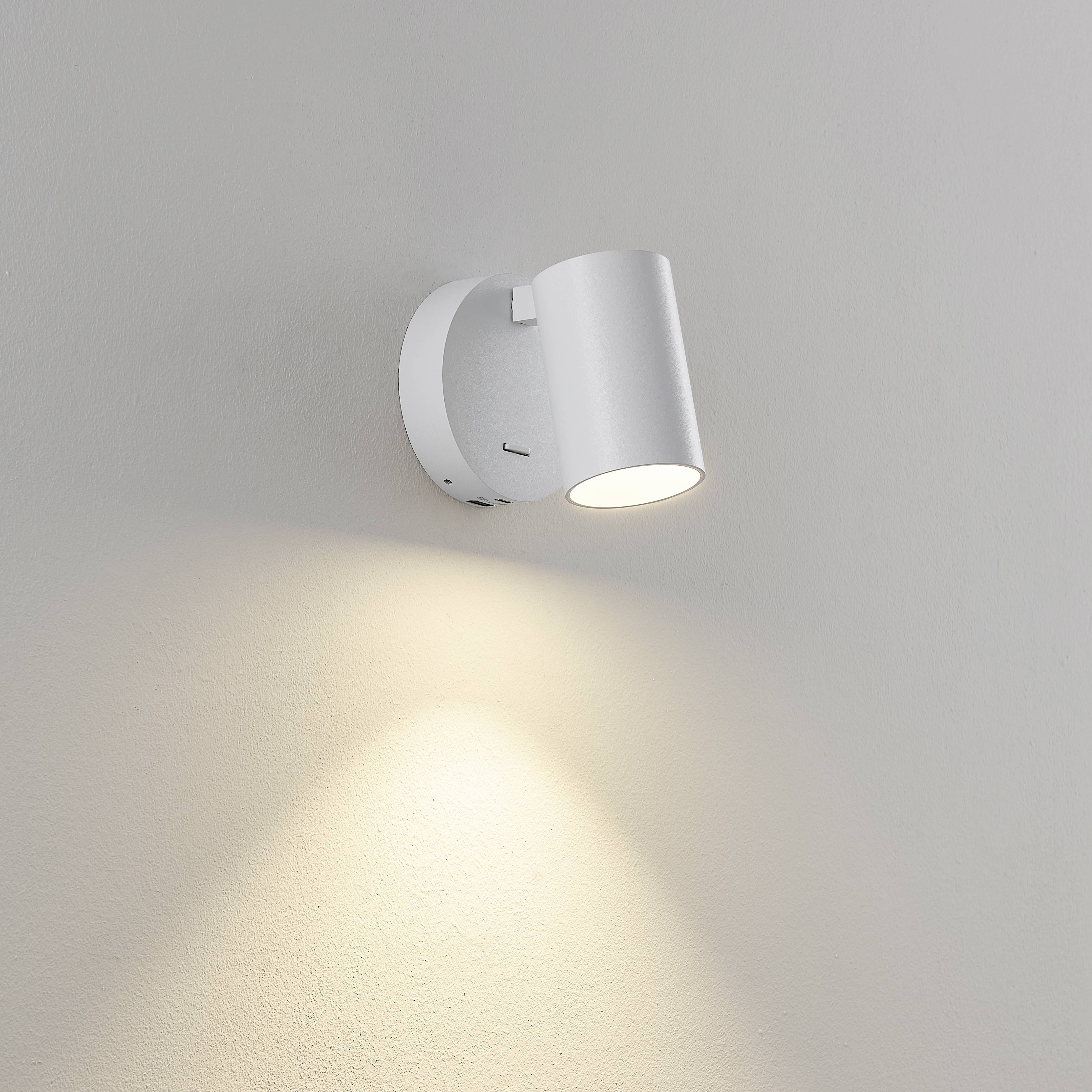 Biała kinkiet emituje ciepłe światło skierowane w dół na gładką, kremową ścianę. Nowoczesny design, idealny jako oświetlenie akcentujące.