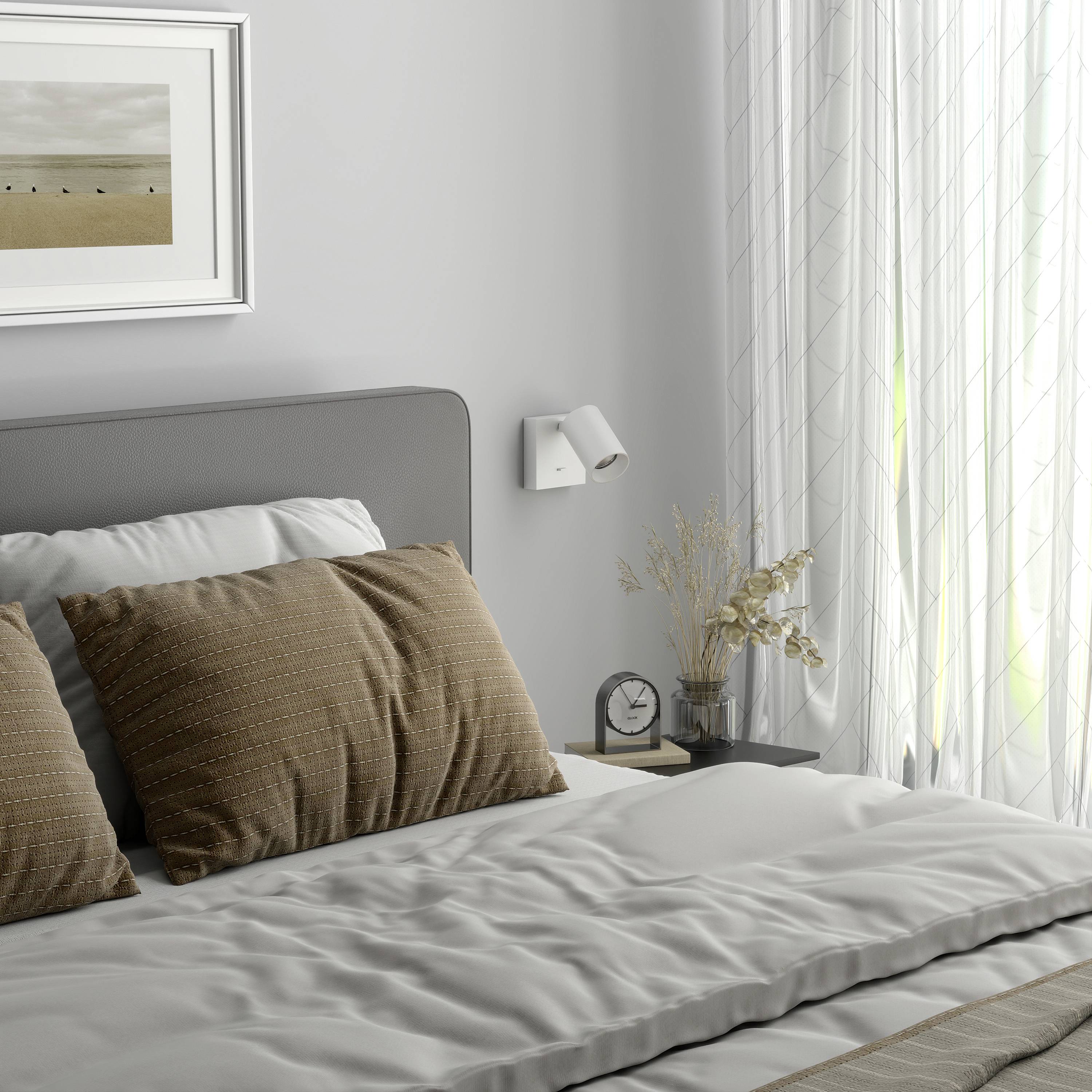 Przytulna sypialnia z szarym łóżkiem, dwoma brązowymi poduszkami, obrazem na ścianie i delikatnym światłem dziennym wpadającym przez zasłony.