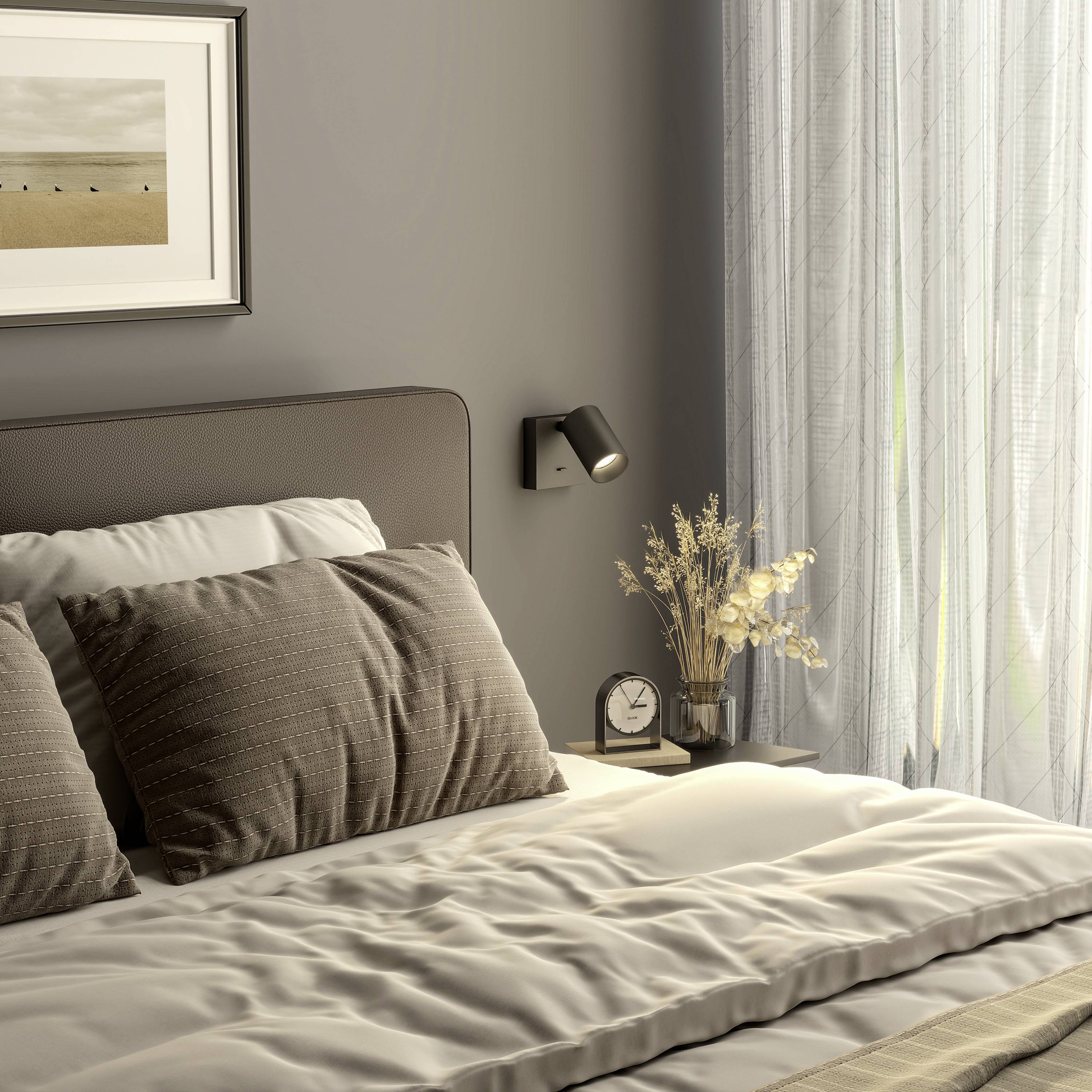 Przytulna sypialnia z szarym łóżkiem, dwoma dużymi poduszkami, stolikiem nocnym z lampą i obrazem na ścienie.