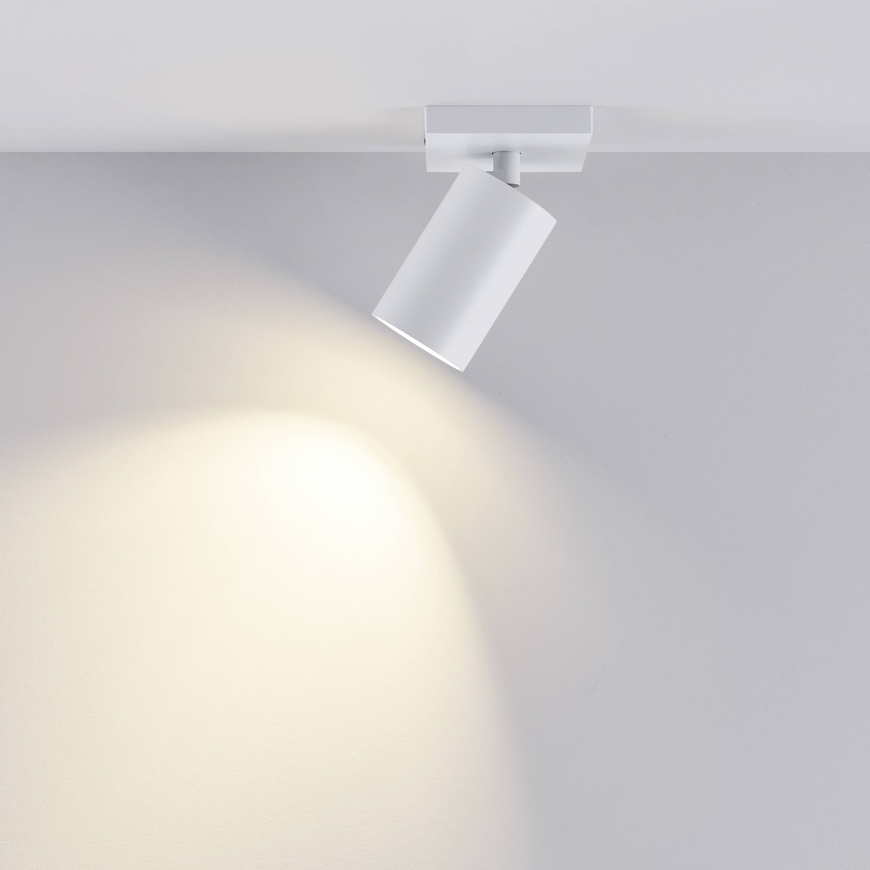 Pojedyncza, nowoczesna biała lampa sufitowa rzuca światło na neutralną, szarą ścianę, tworząc czysty, skoncentrowany stożek świetlny.