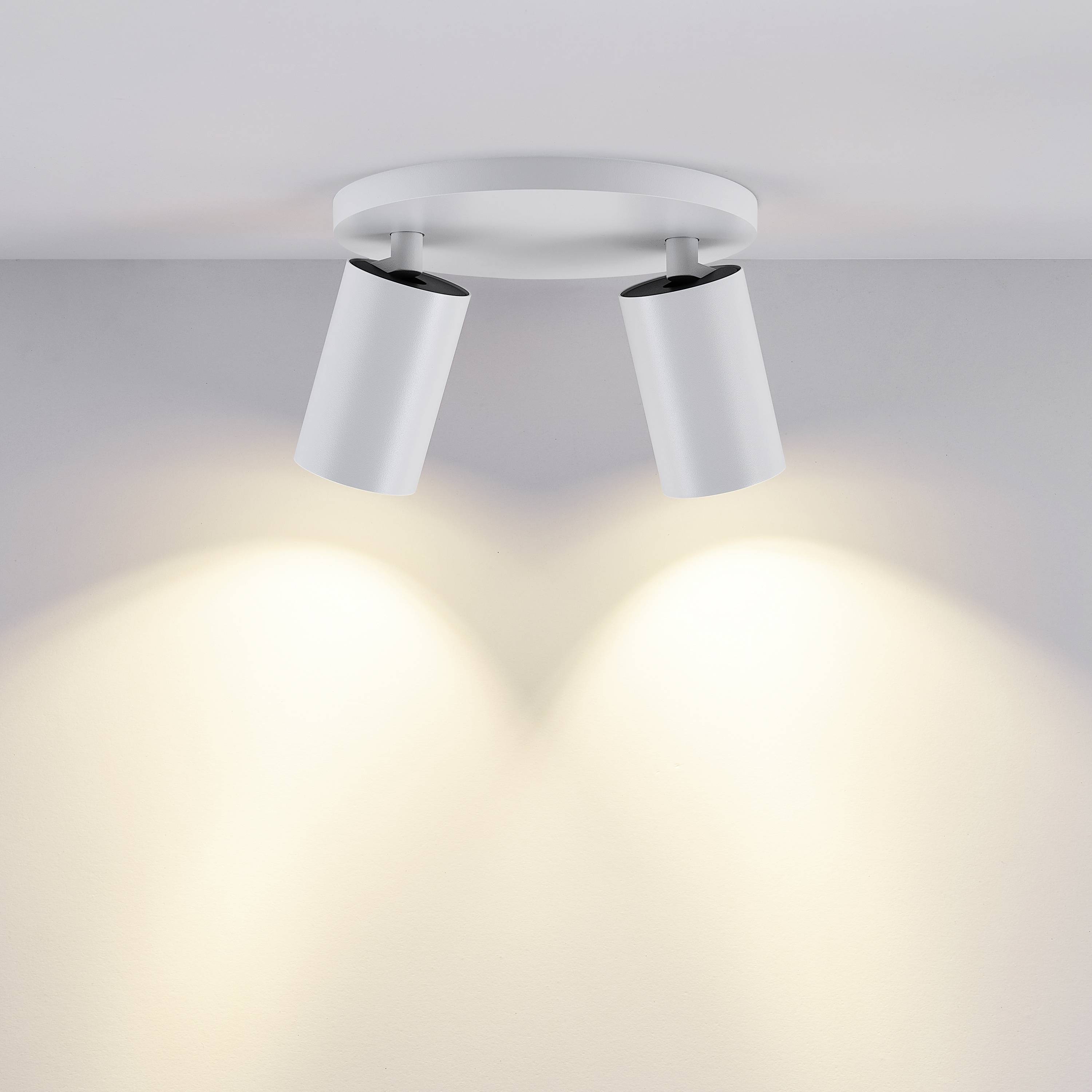 Lampa sufitowa z dwoma białymi, cylindrycznymi reflektorami, które rzucają miękkie, żółte światło na ścianę.