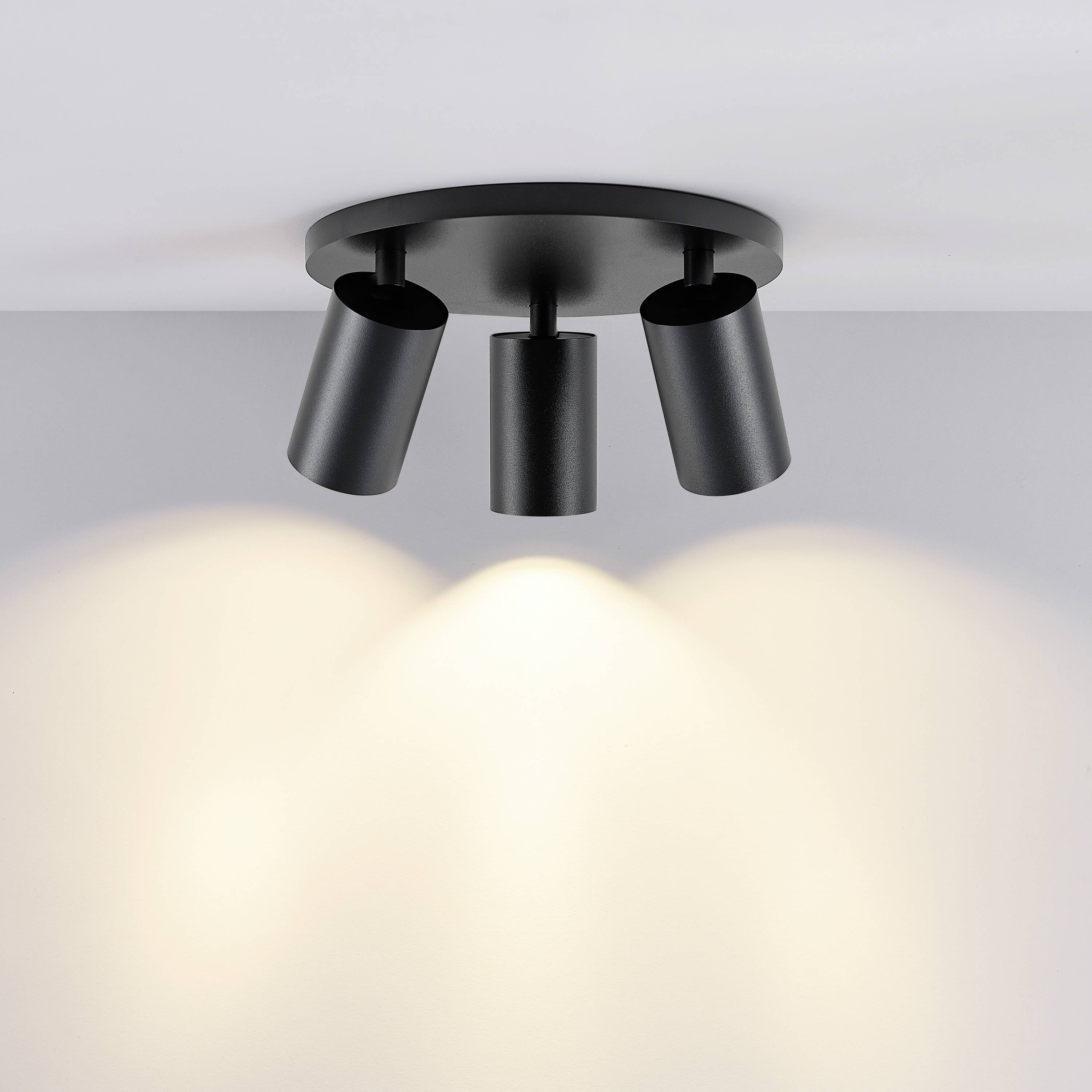 Lampa sufitowa z trzema regulowanymi reflektorami w matowej czerni, rzucająca miękkie, ciepłe światło na powierzchnię poniżej.
