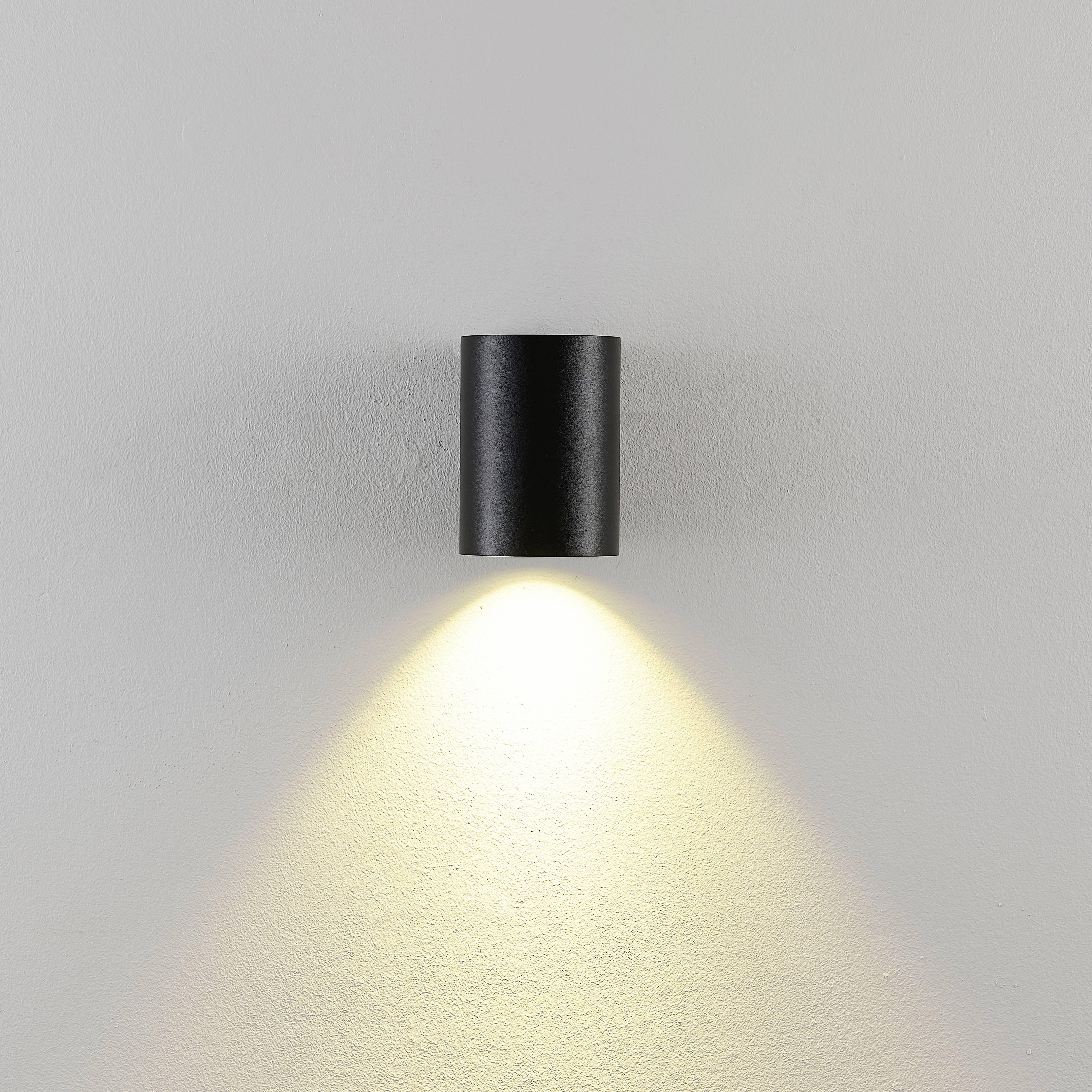 Czarna lampa ścienna rzuca ciepłe światło na białą ścianę.
