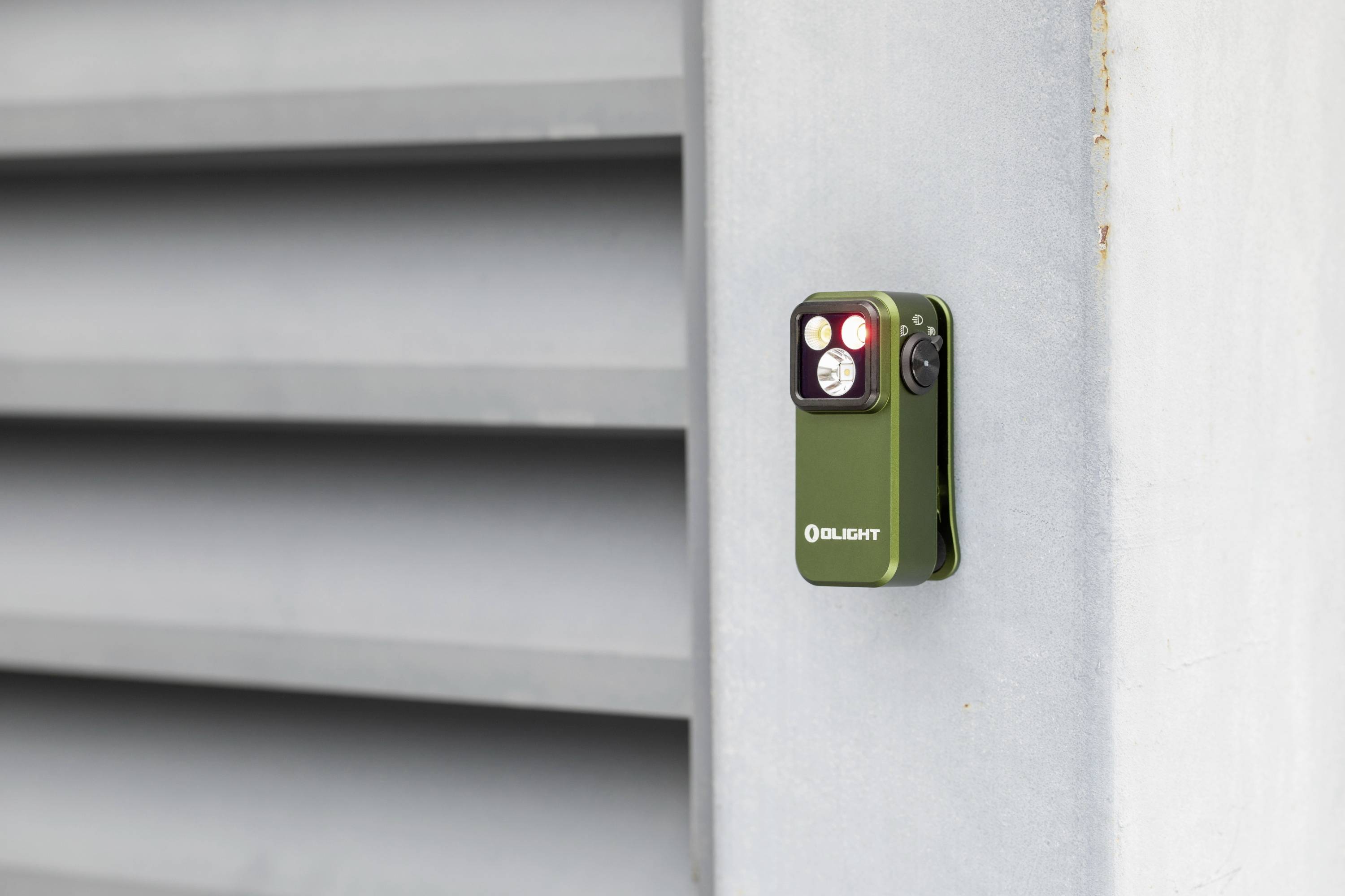 Mała, zielona latarka z napisem 'Olight' jest przymocowana do metalowej, prążkowanej powierzchni.