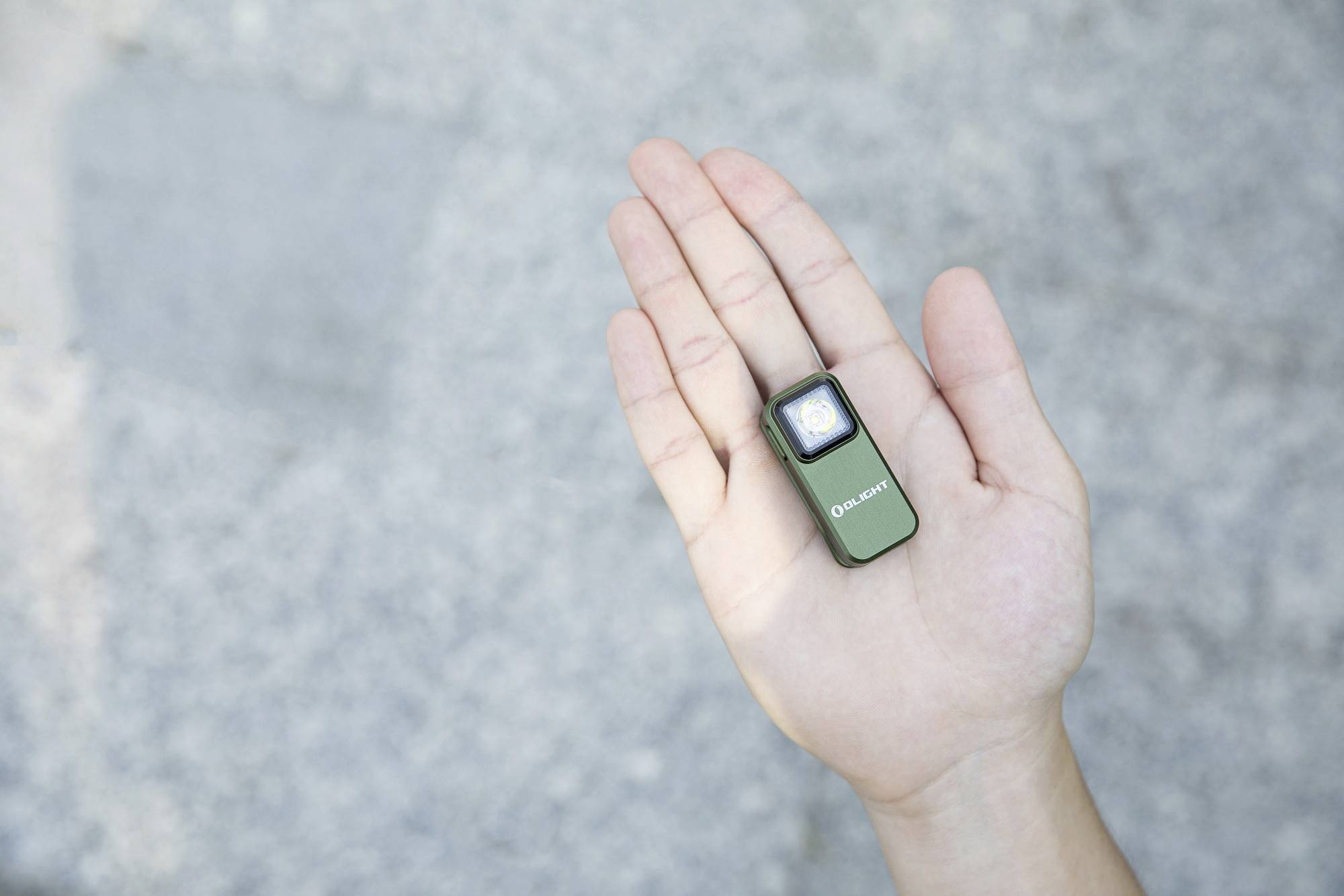 Osoba trzyma małą zieloną latarkę na dłoni, która jest włączona. Tło jest szare i rozmyte.