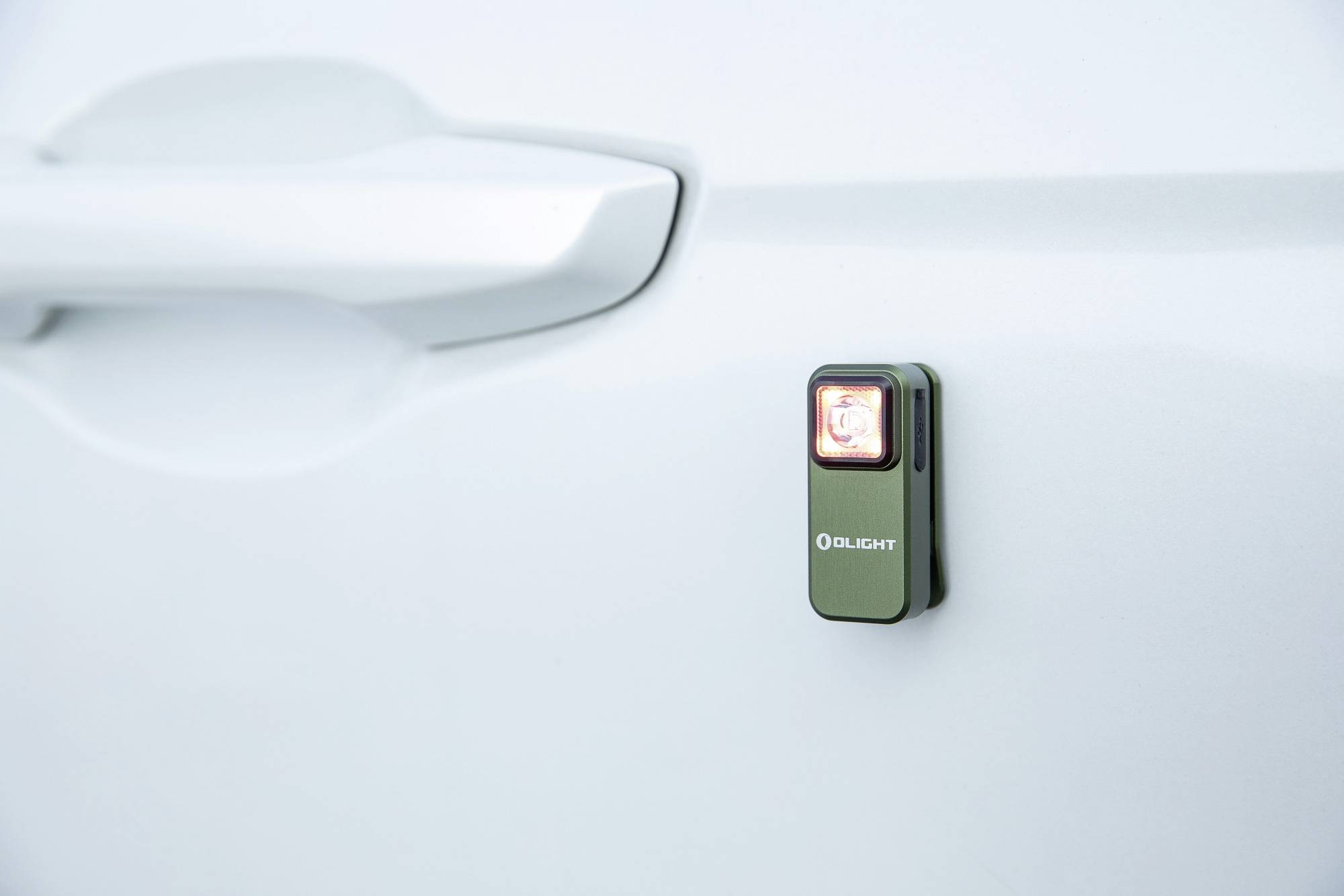 Mała, zielona lampka marki 'Olight' jest przymocowana do drzwi samochodu i świeci na czerwono.