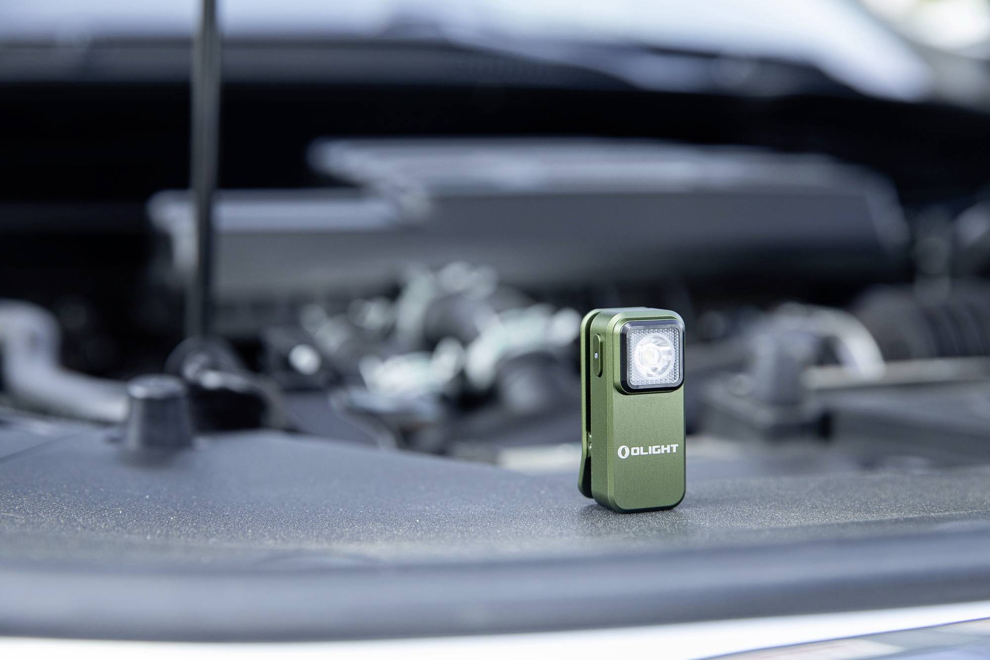 Mała, zielona latarka stoi na silniku samochodu. W tle widać rozmazany przedział silnika.