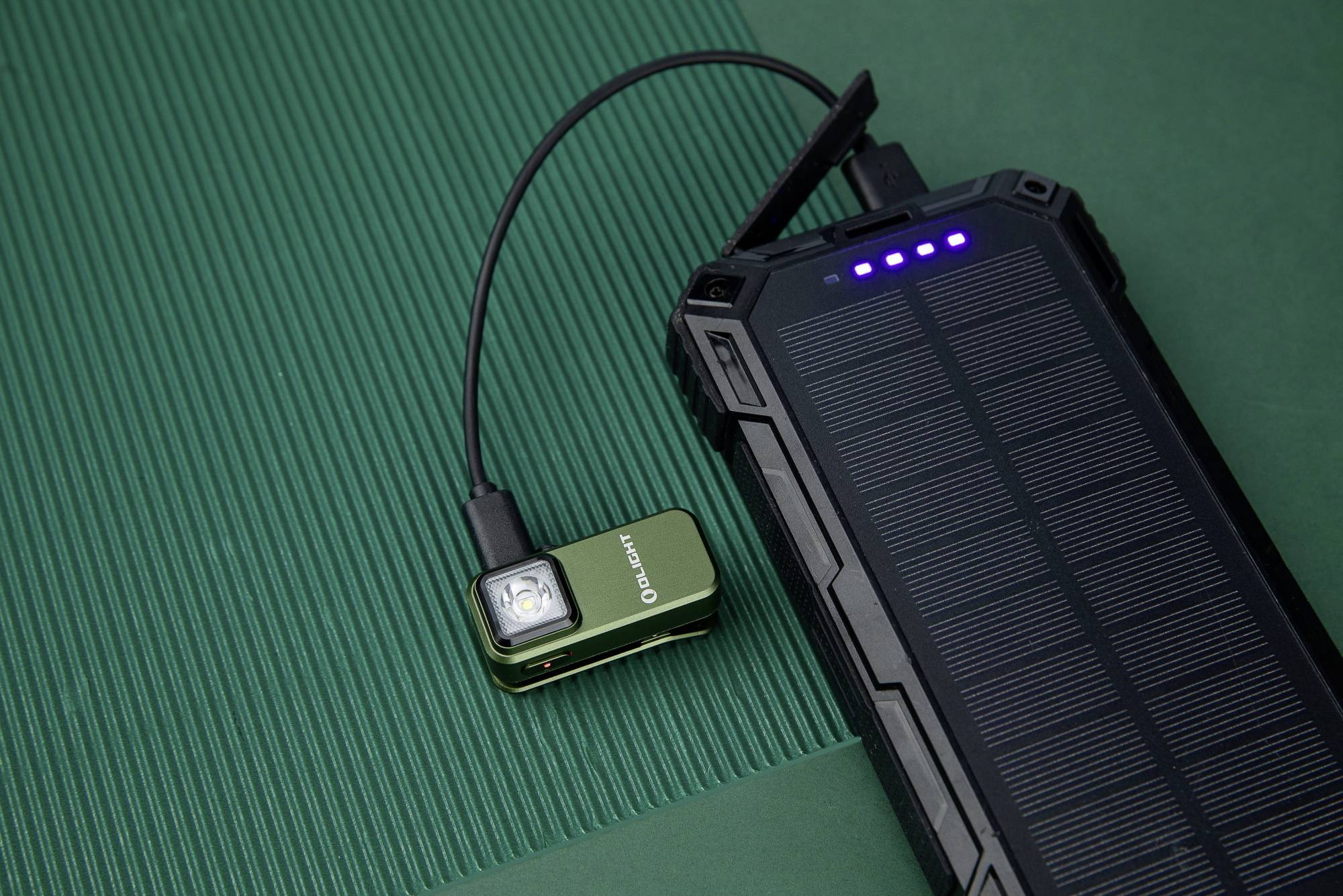 Duży, czarny solarny ładowarka z czterema niebieskimi światłami ładuje małą, zieloną latarkę za pomocą kabla.