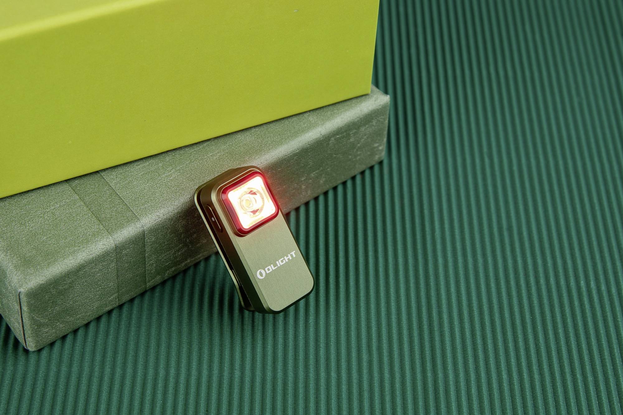 Mała czarna latarka z czerwonym światłem świeci na zielonym, prążkowanym podłożu obok żółtego kartonu.